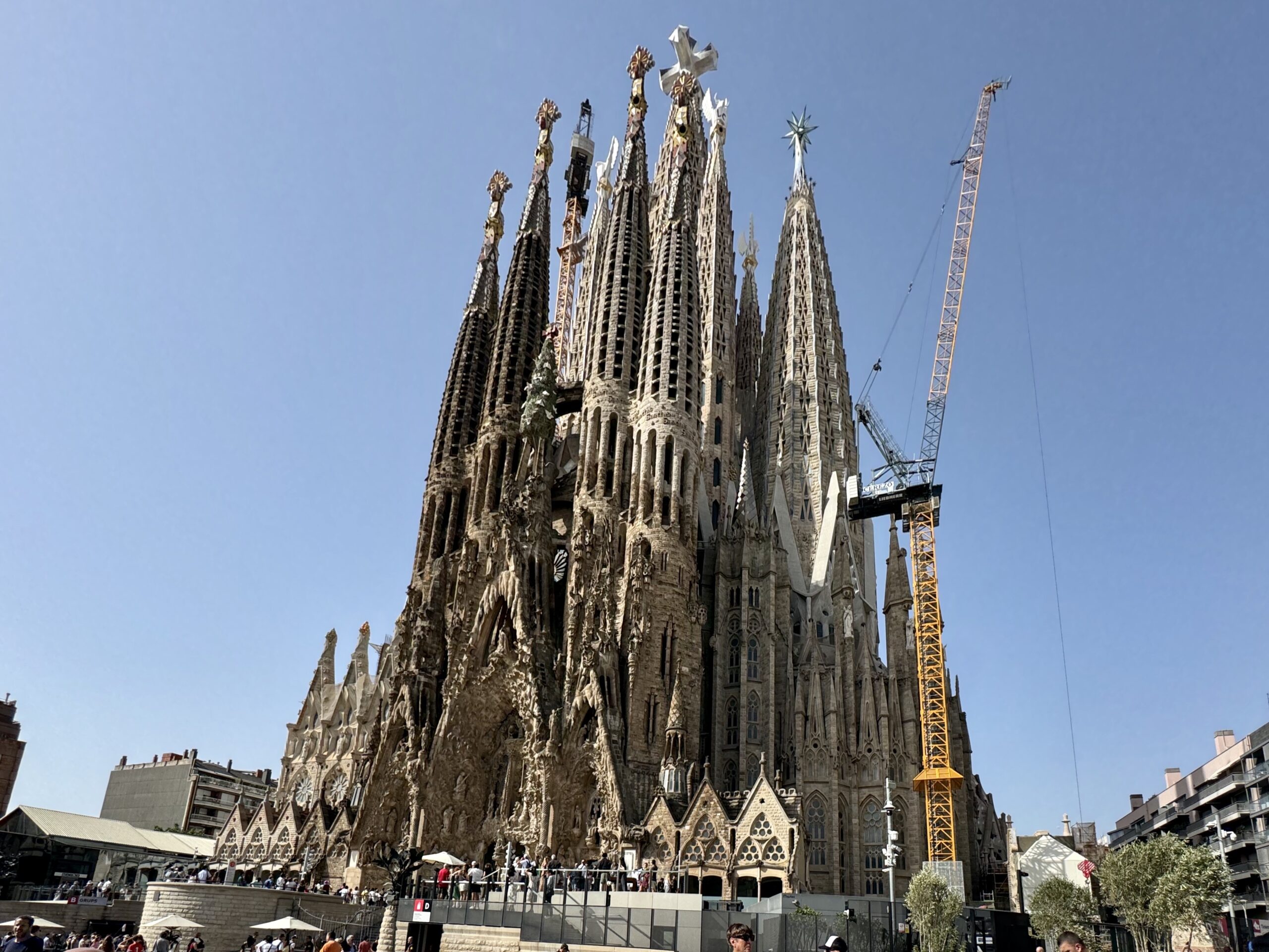 Sagrada Família towers in Barcelona with a construction crane against a blue sky, visitors at the base.