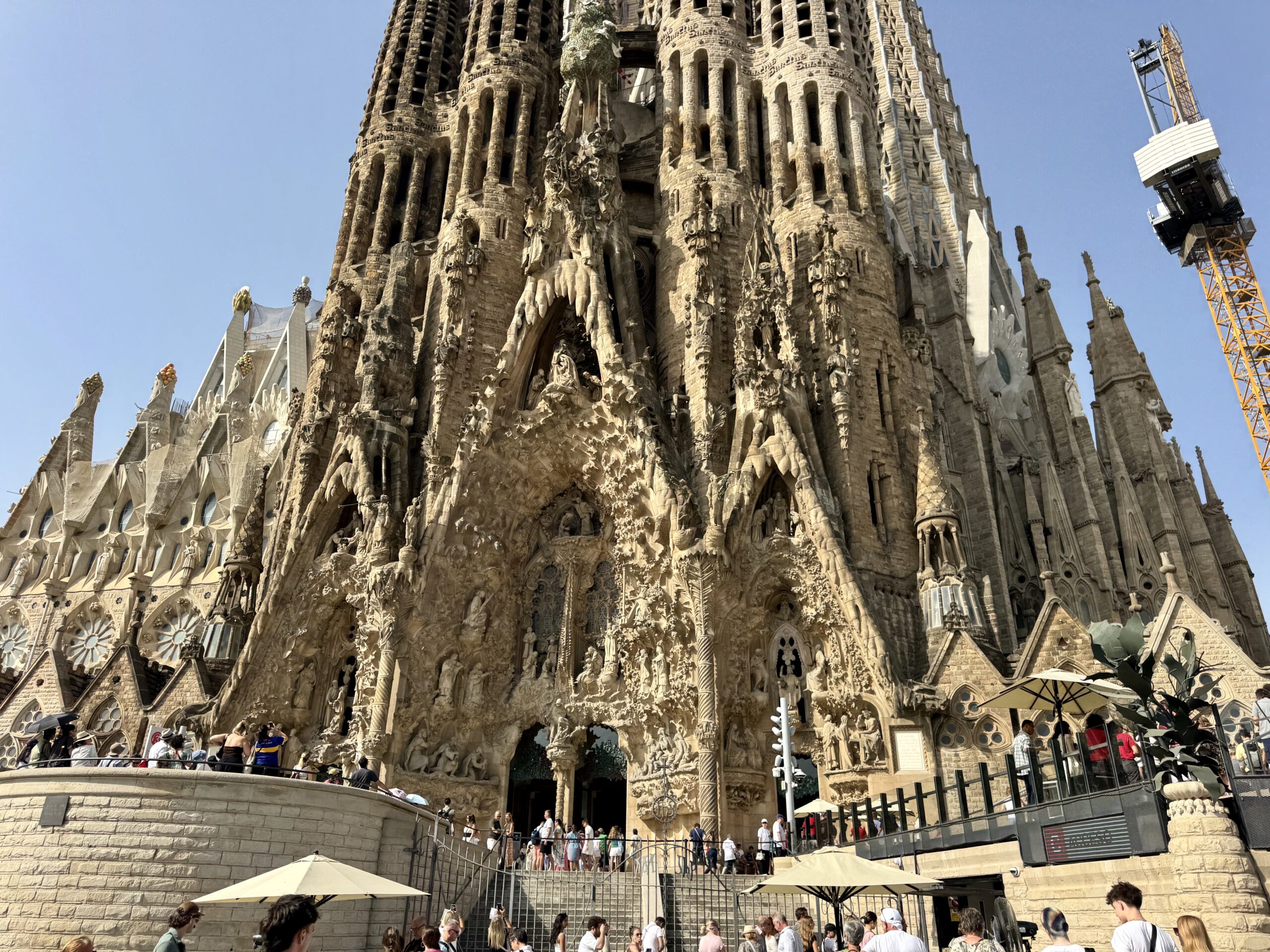 Tourists stand on a stone terrace in front of La Sagrada Família's intricate façade and towers under a clear blue sky.