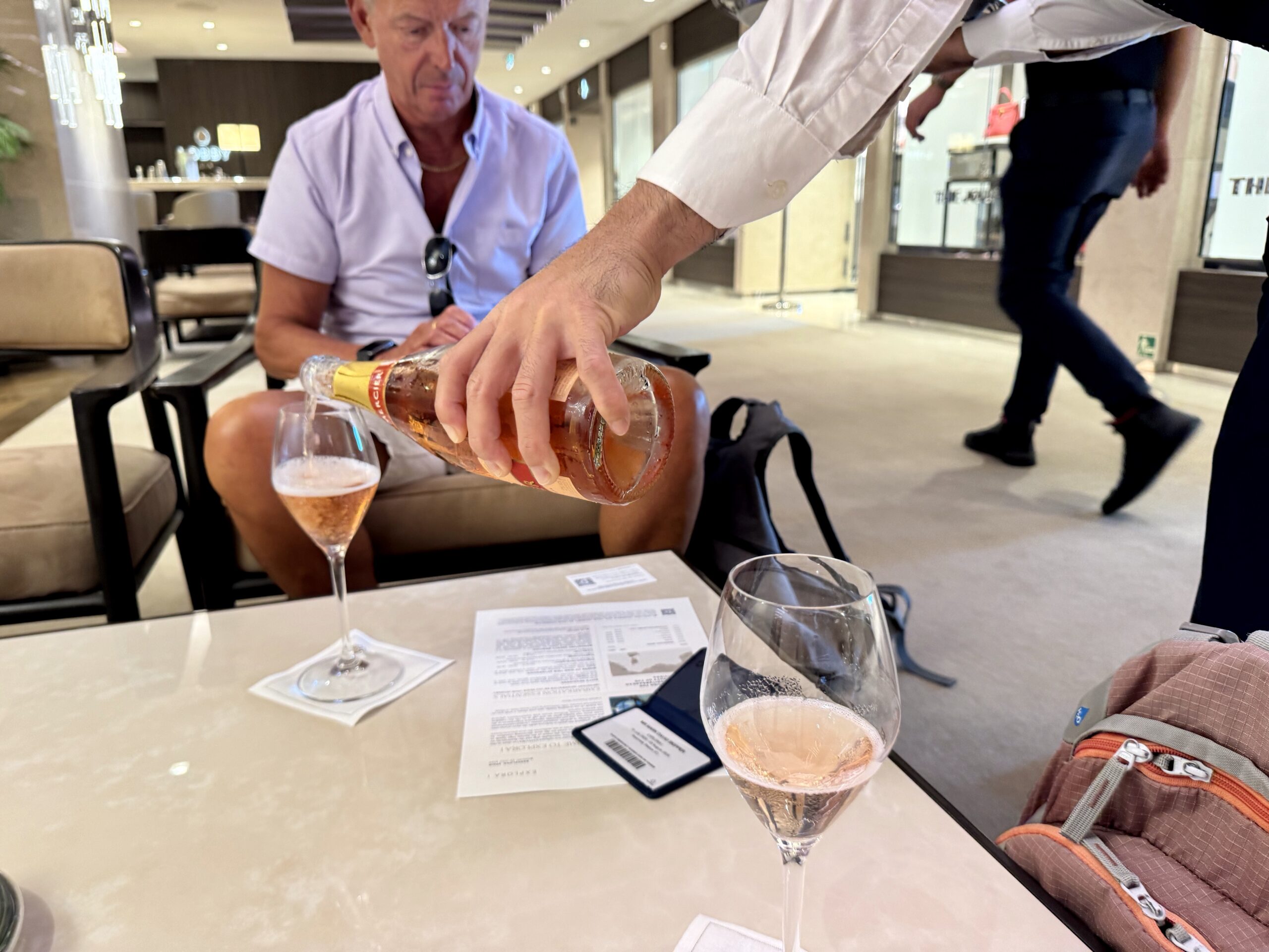 Man pours rosé wine from a bottle into a glass at a dining table indoors; another person sits nearby in a bright lounge.
