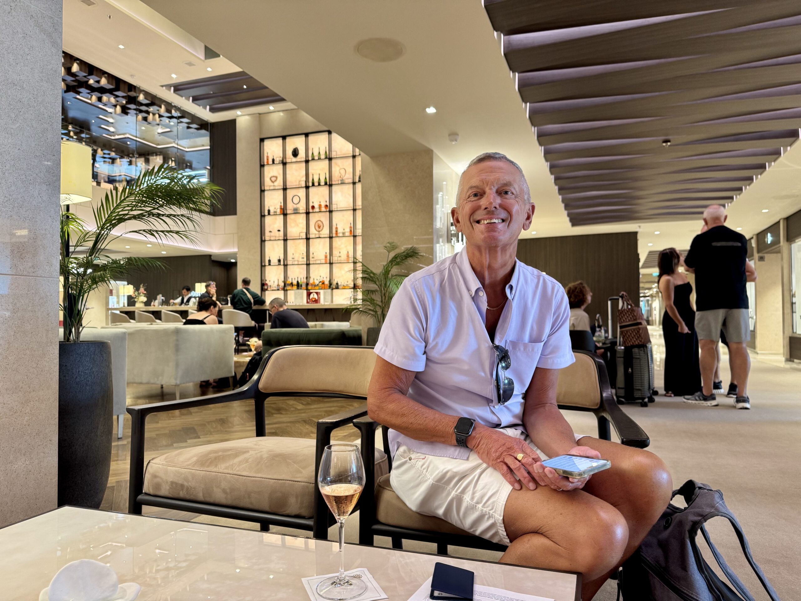 Smiling man in a light shirt sits in a modern hotel lobby, a glass of wine on the table in front of him and sunglasses hanging from his shirt.