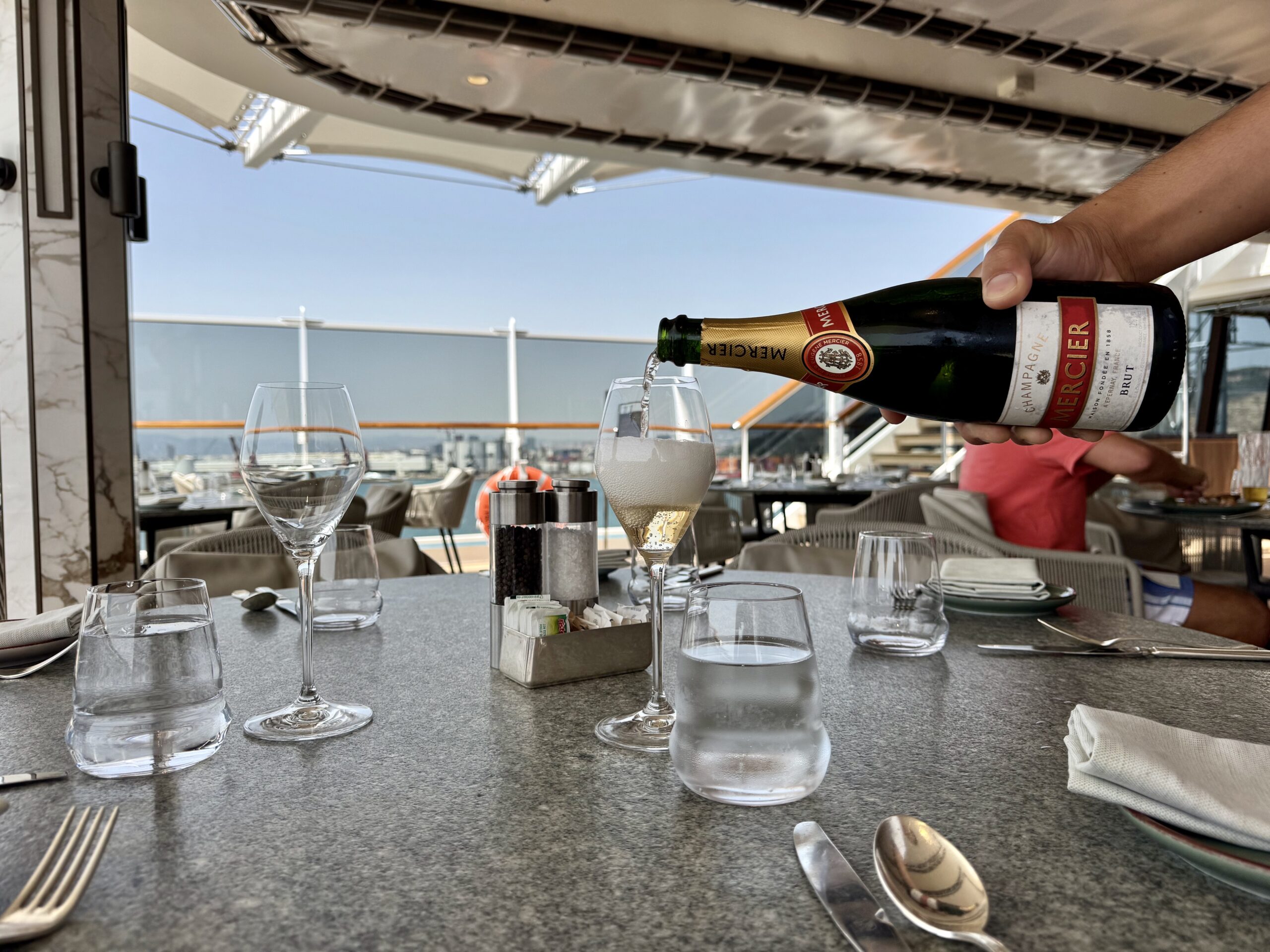 Person pouring champagne from a bottle into a stemmed glass at an outdoor restaurant table on a sunny day.