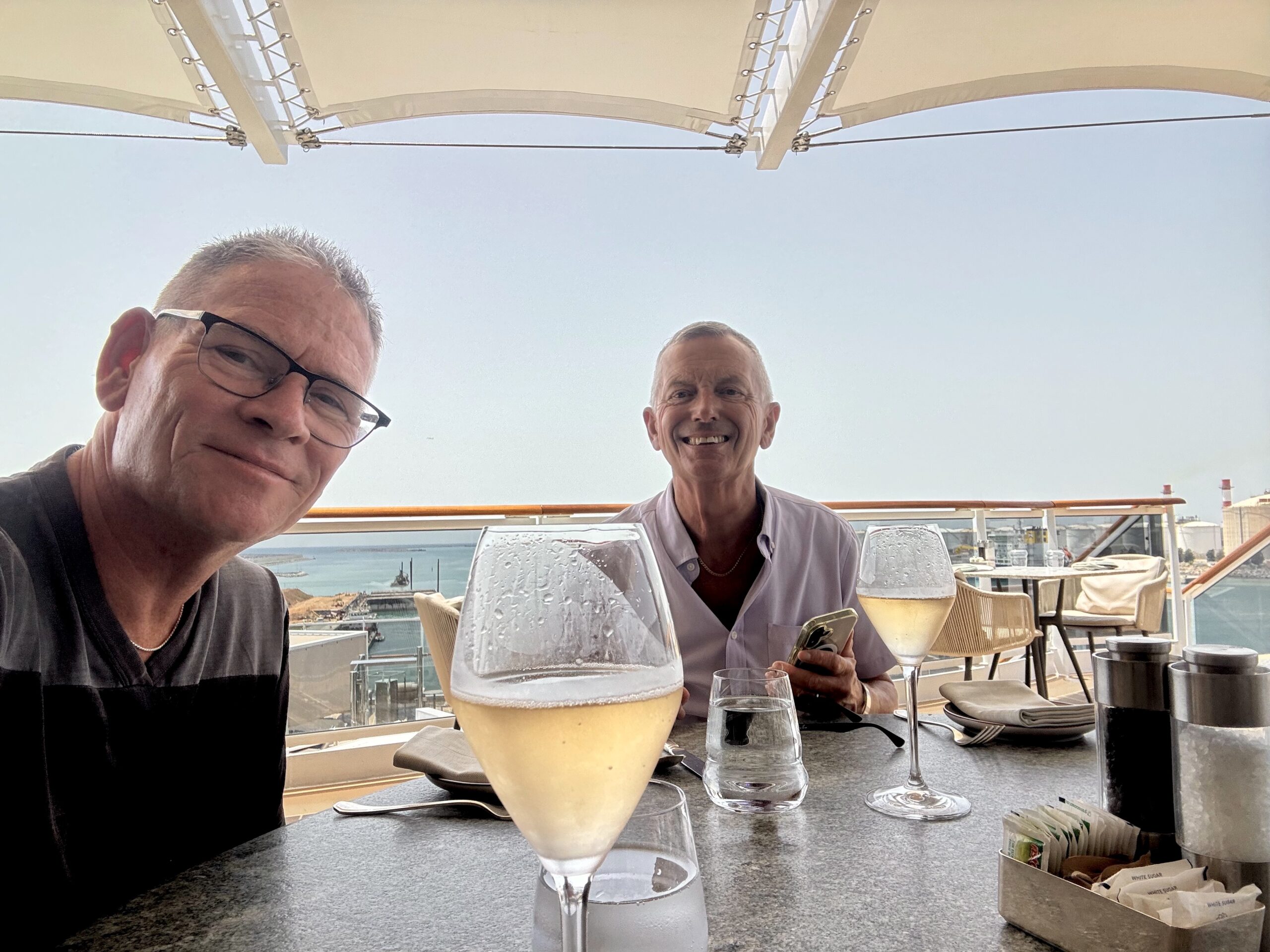 Two men smiling at an outdoor waterfront restaurant, with frosty cocktails on the table and a blue sea in the background.