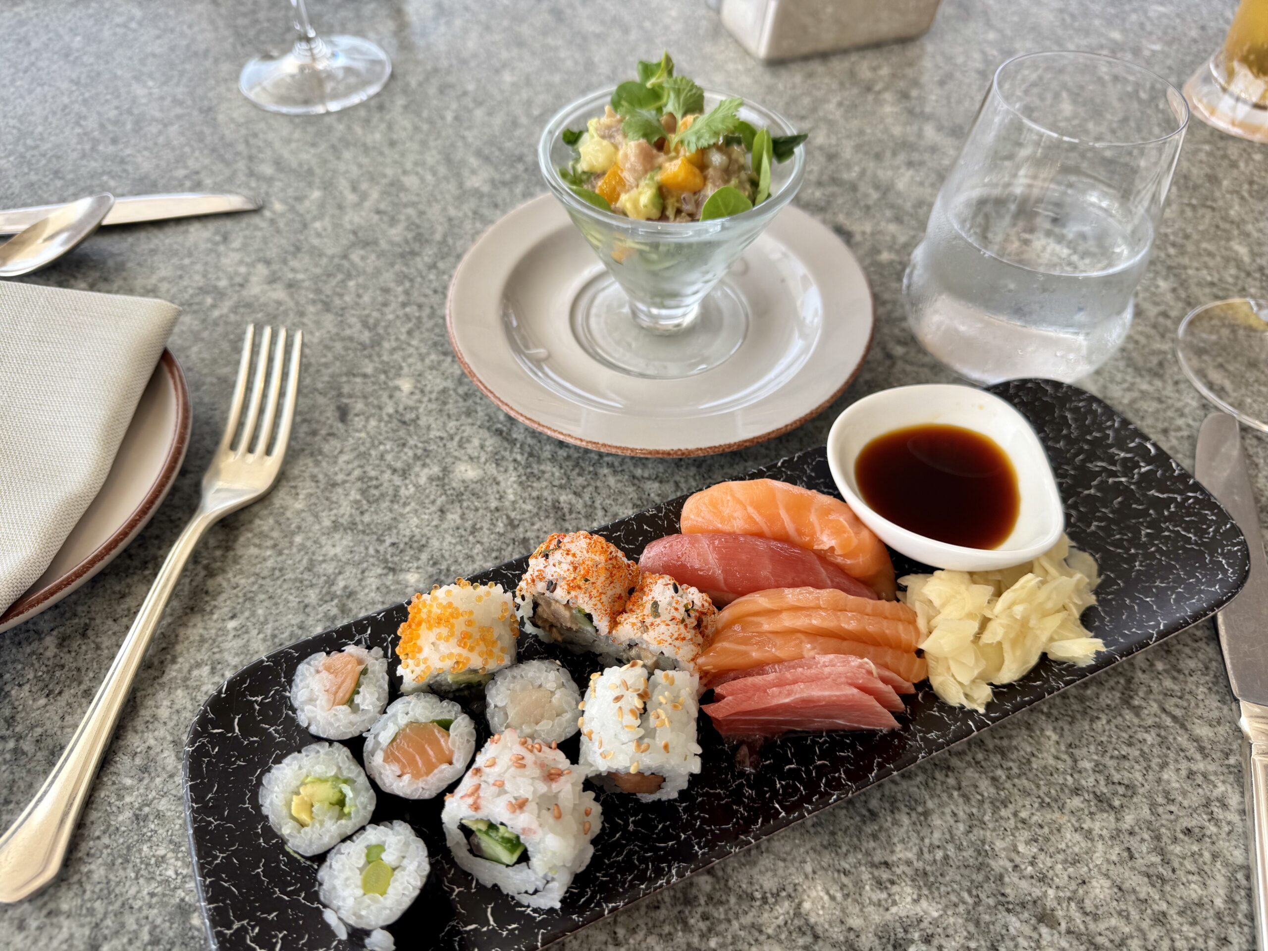 Assorted sushi platter with nigiri and rolls on a dark marble board, soy sauce and pickled ginger nearby, glassware in the background