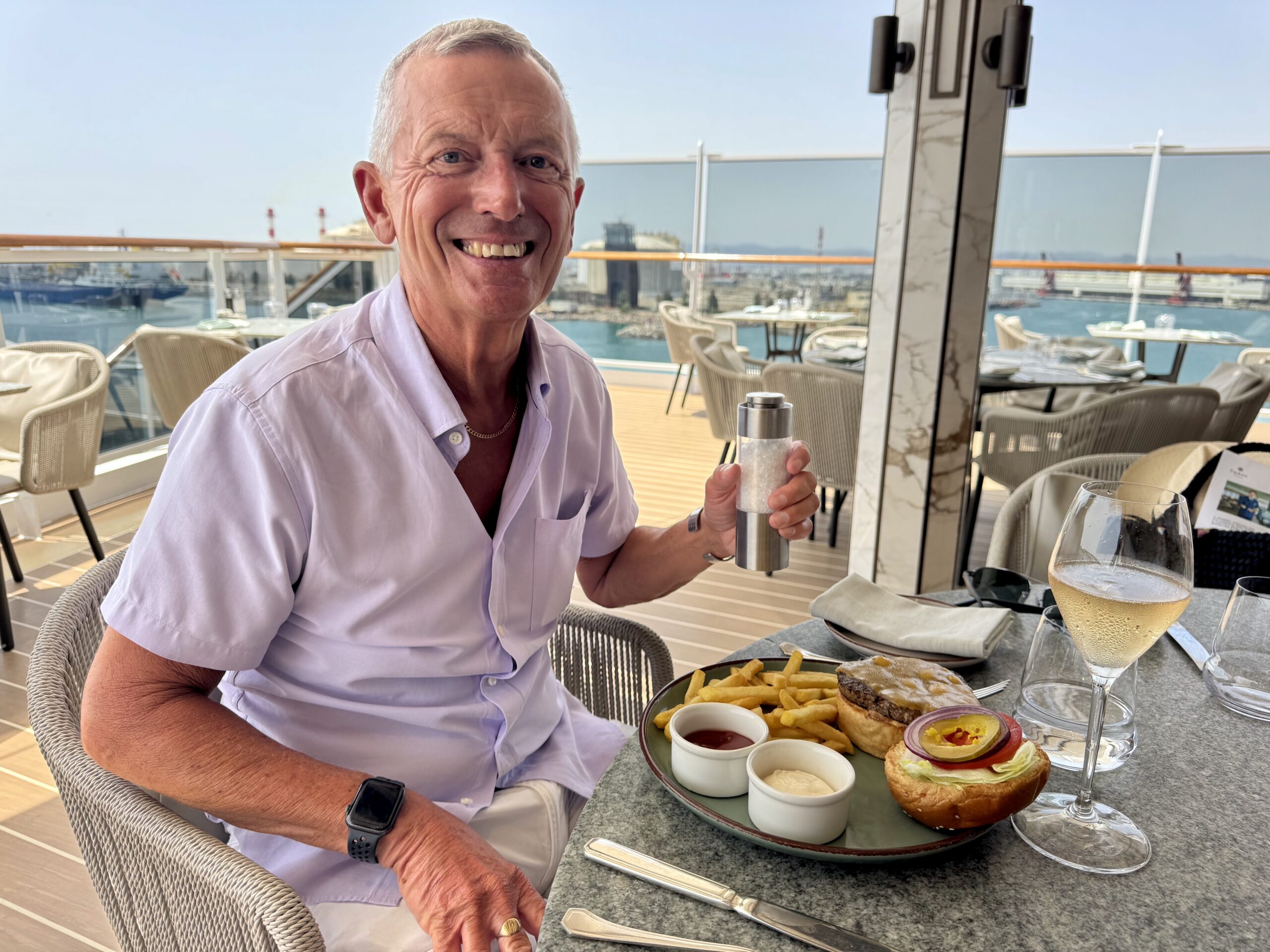Smiling older man in a light shirt sits at an outdoor harbor restaurant, holding a salt grinder toward the camera.