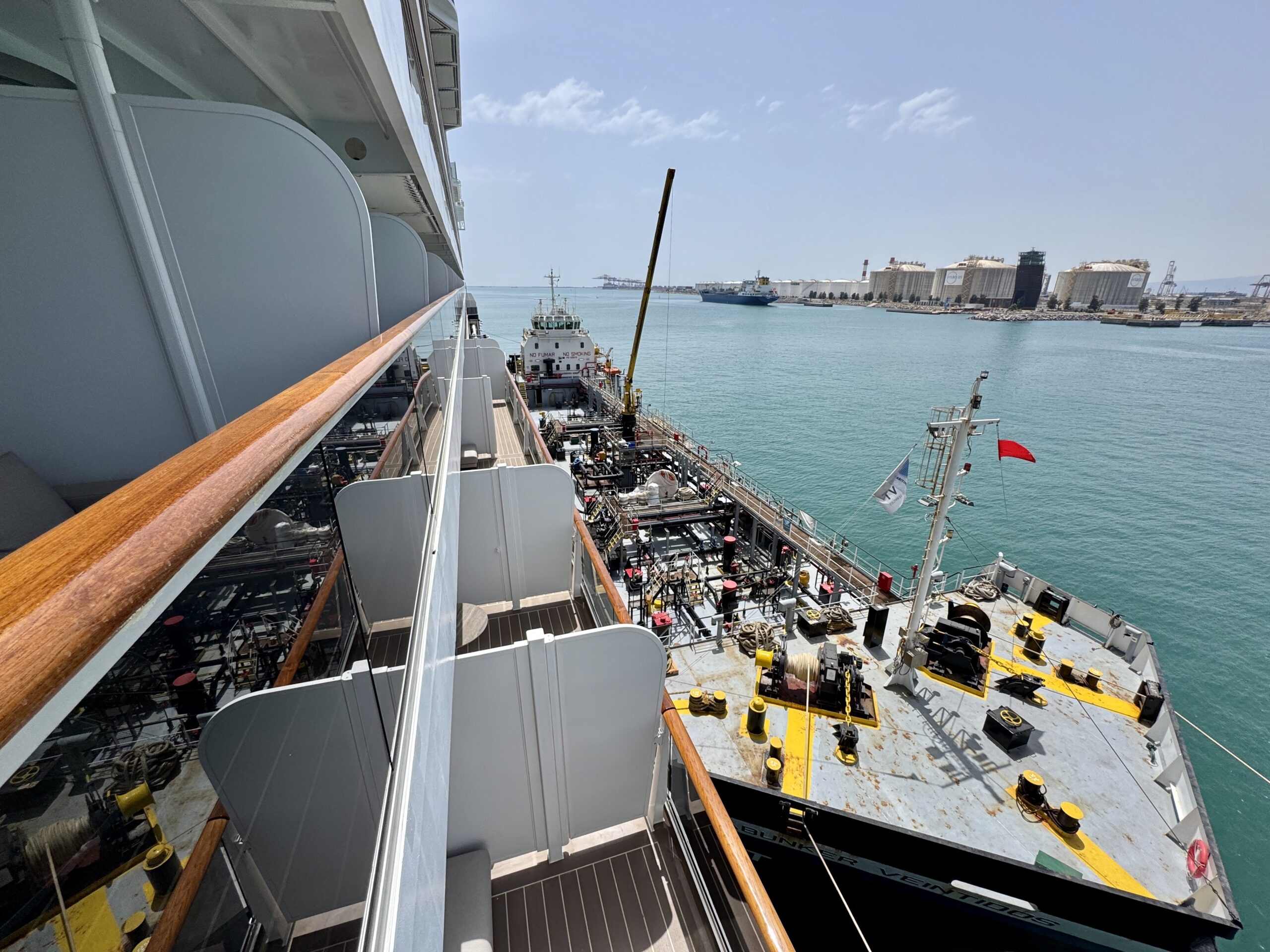 View from a ship deck looking toward the harbor with turquoise water and industrial port buildings in the distance, metal railing and deck equipment in the foreground.