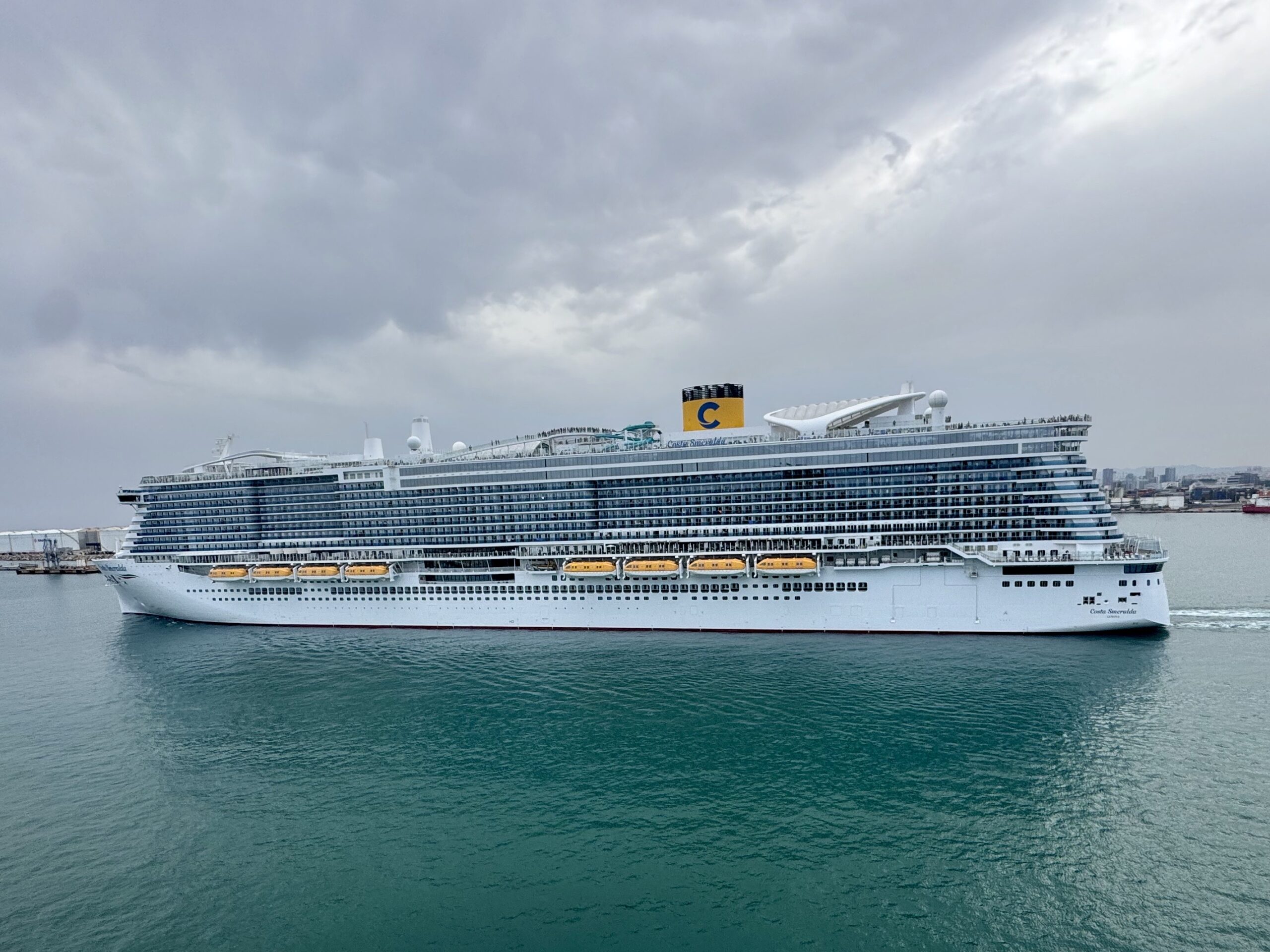Large white cruise ship with many decks on teal water under a cloudy gray sky, lifeboats visible along the side.
