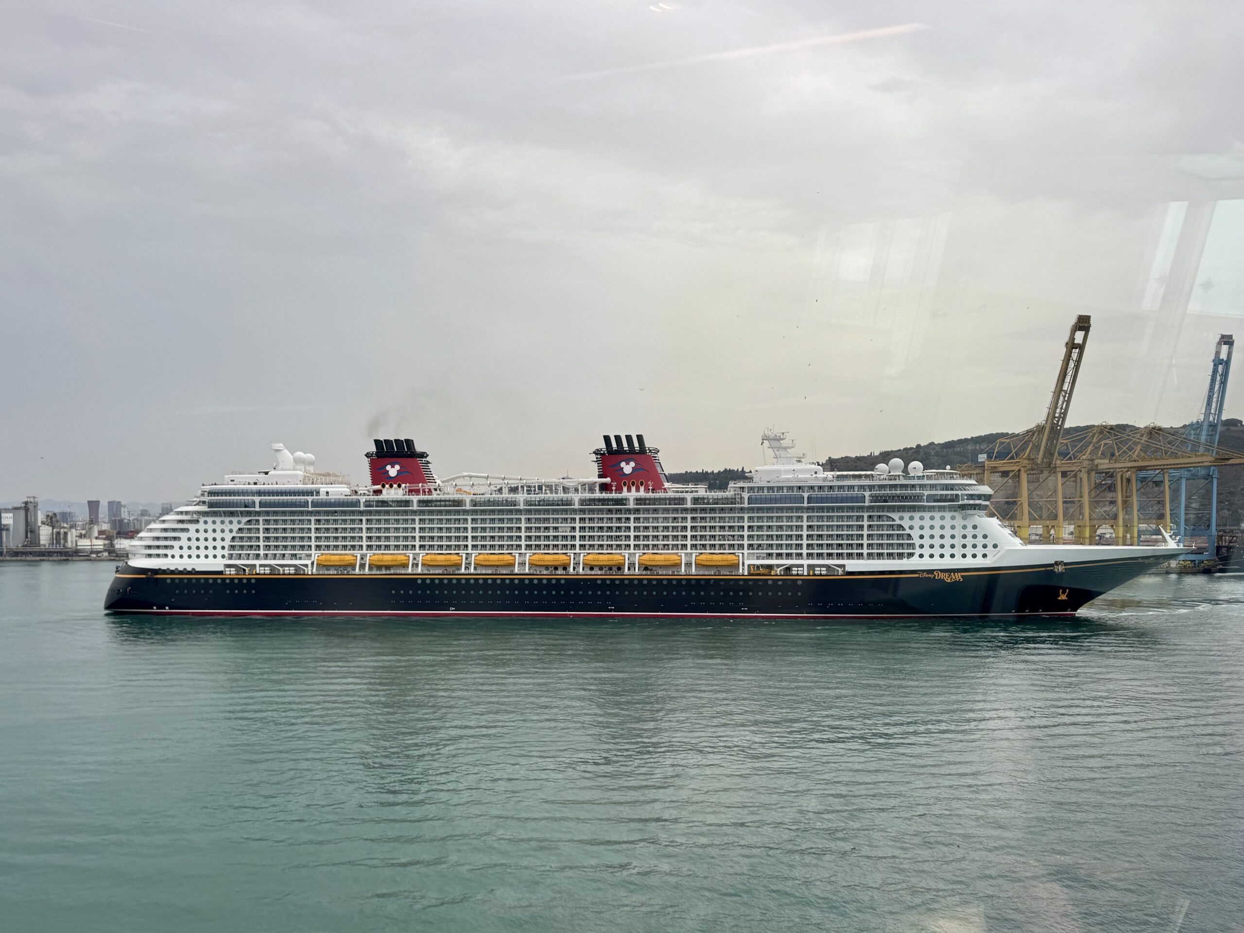 Large cruise ship docked at a harbor with cranes on the right and calm water in the foreground