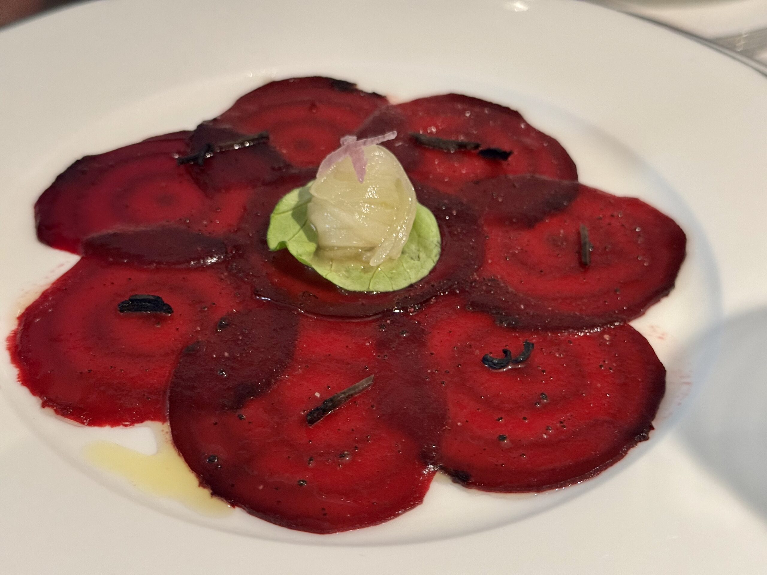Beet carpaccio arranged on a white plate with a small green leaf garnish and onion shard on top.