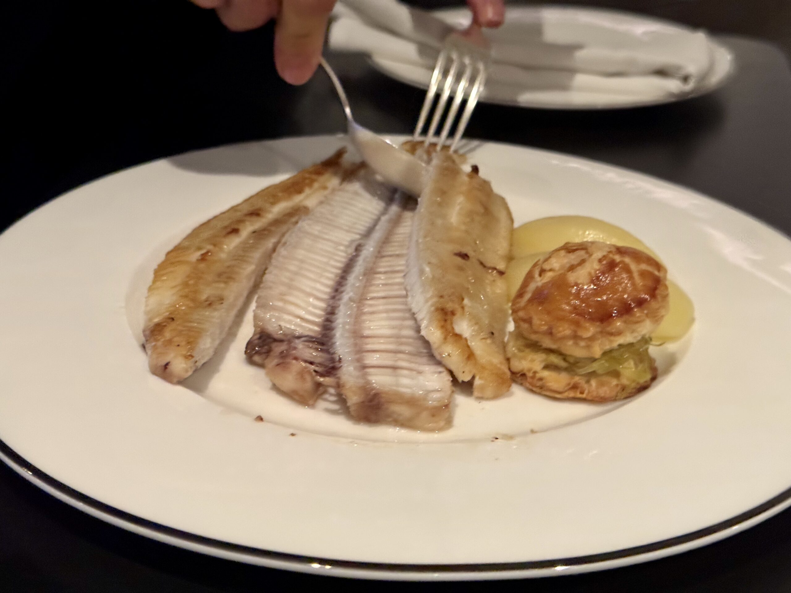 Plate with sliced roasted fish fillet, two potato dumplings, and a roasted potato on a white dish; fork resting above the fish.