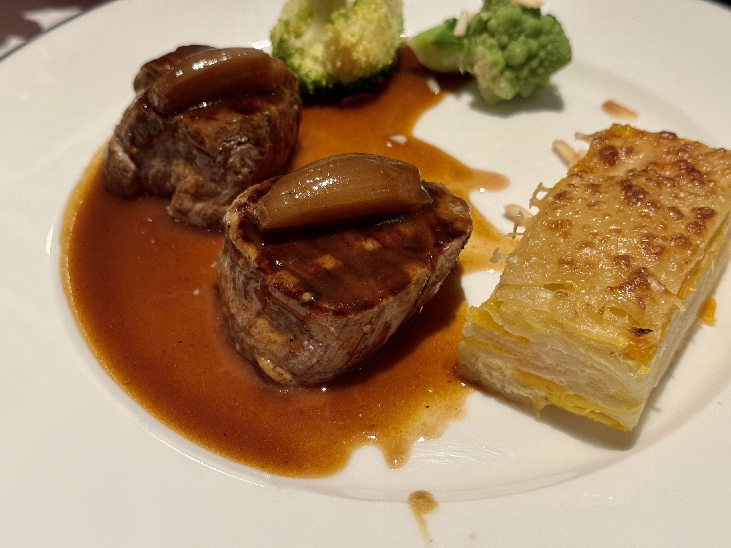 Two beef medallions in brown sauce with onion, beside broccoli and potato gratin on a white plate.