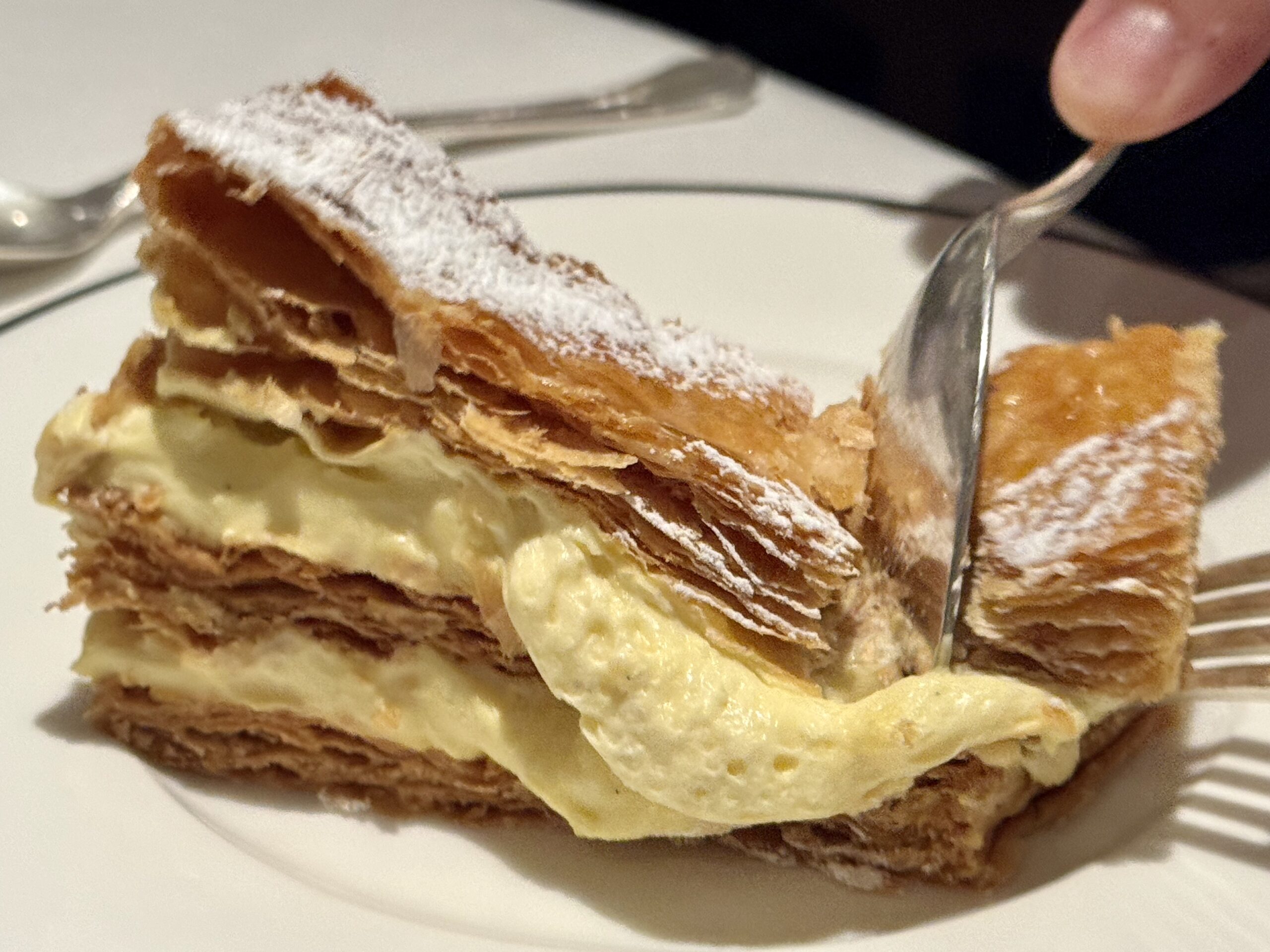 Close-up of a cream-filled puff pastry slice with flaky layers and a dusting of powdered sugar on a white plate.