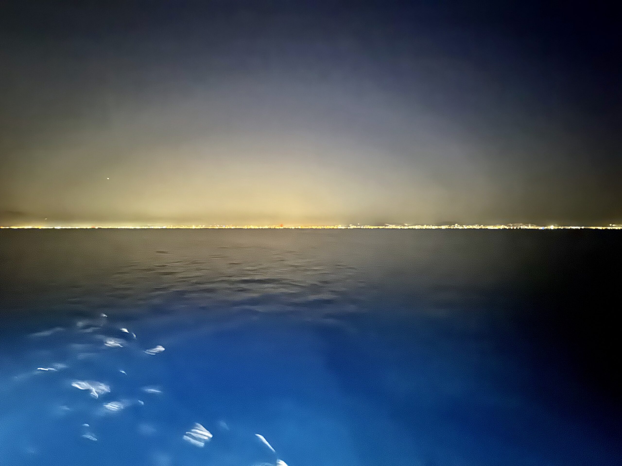 Night seascape with a distant line of lights along the horizon and a dark blue sky above the calm water.