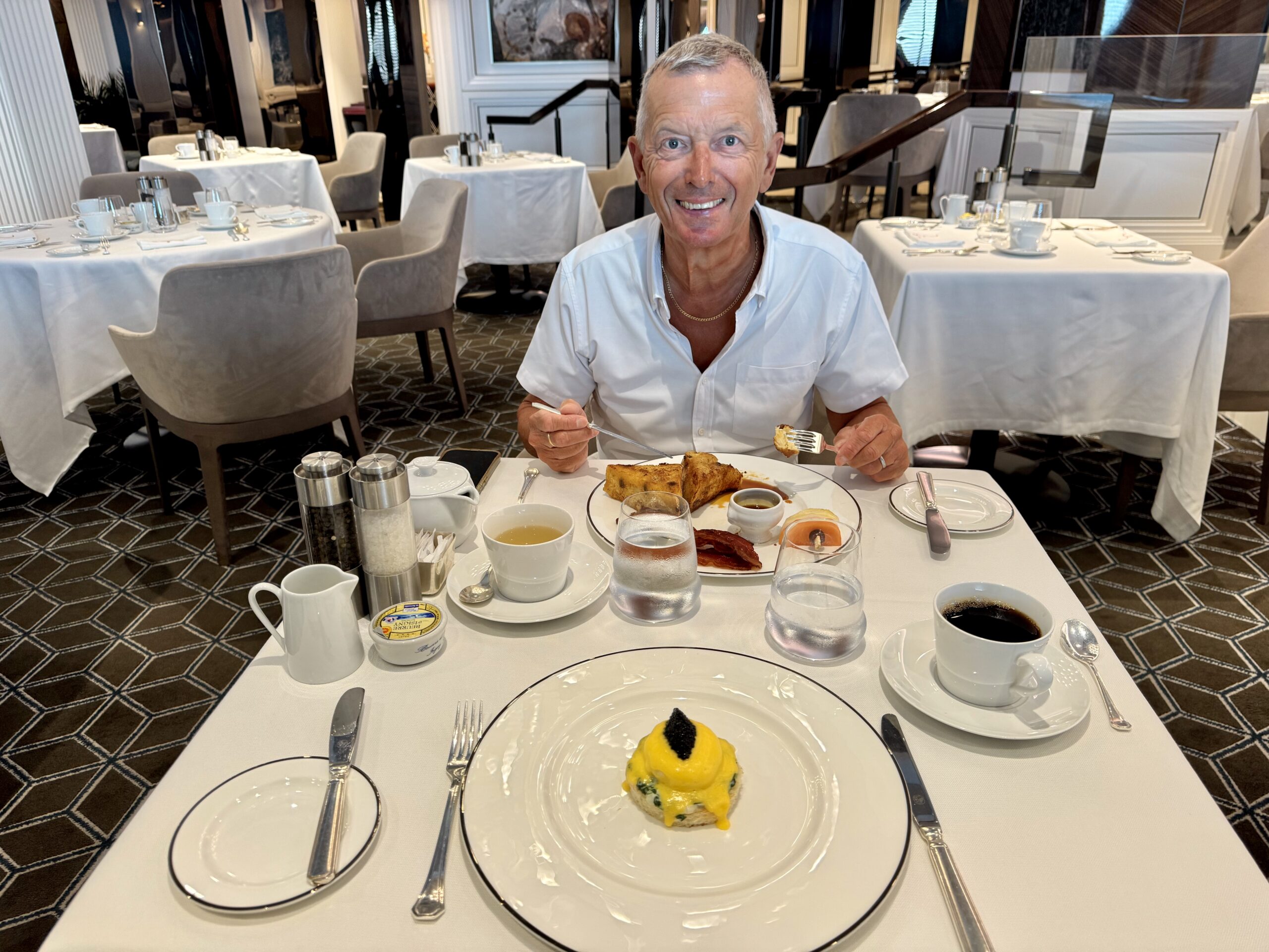 Smiling man dining alone at a white-tablecloth restaurant table, several plates and cups set around him, enjoying a meal.