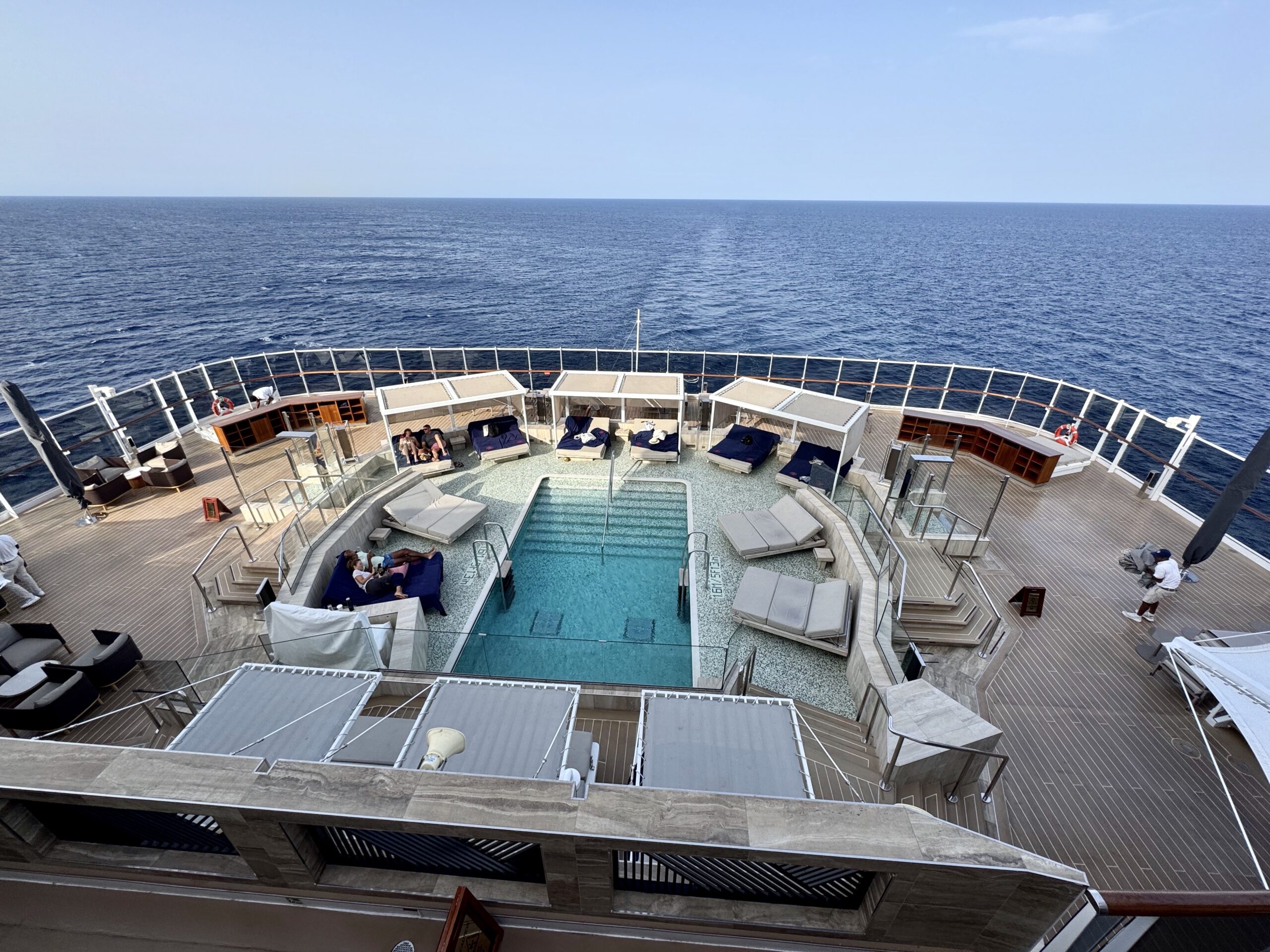 Top deck of a cruise ship with a central pool, white railings, and loungers overlooking the open ocean.