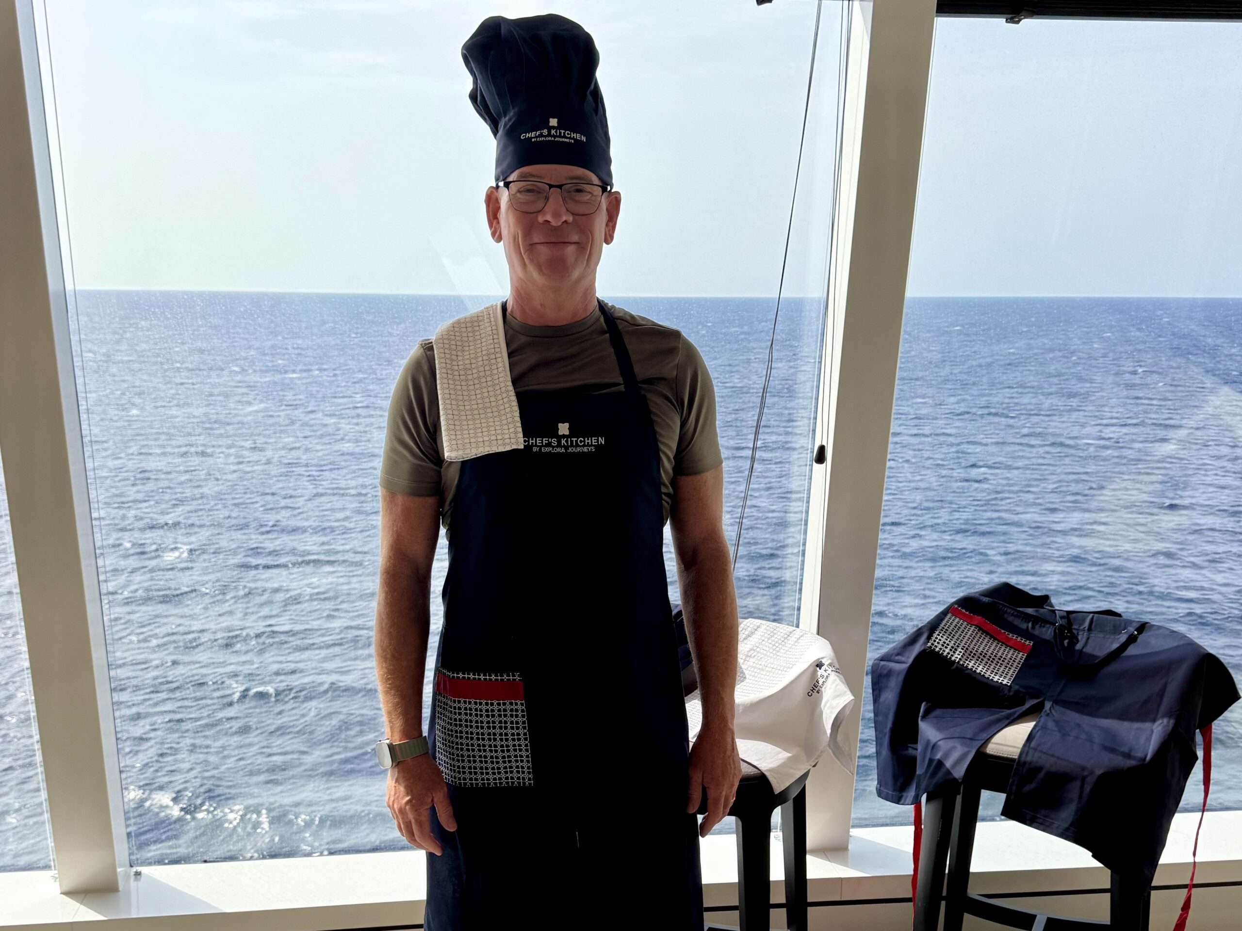 Smiling man wearing a chef hat and apron with a towel over his shoulder, standing in a ship kitchen area with the ocean visible through large windows behind him.