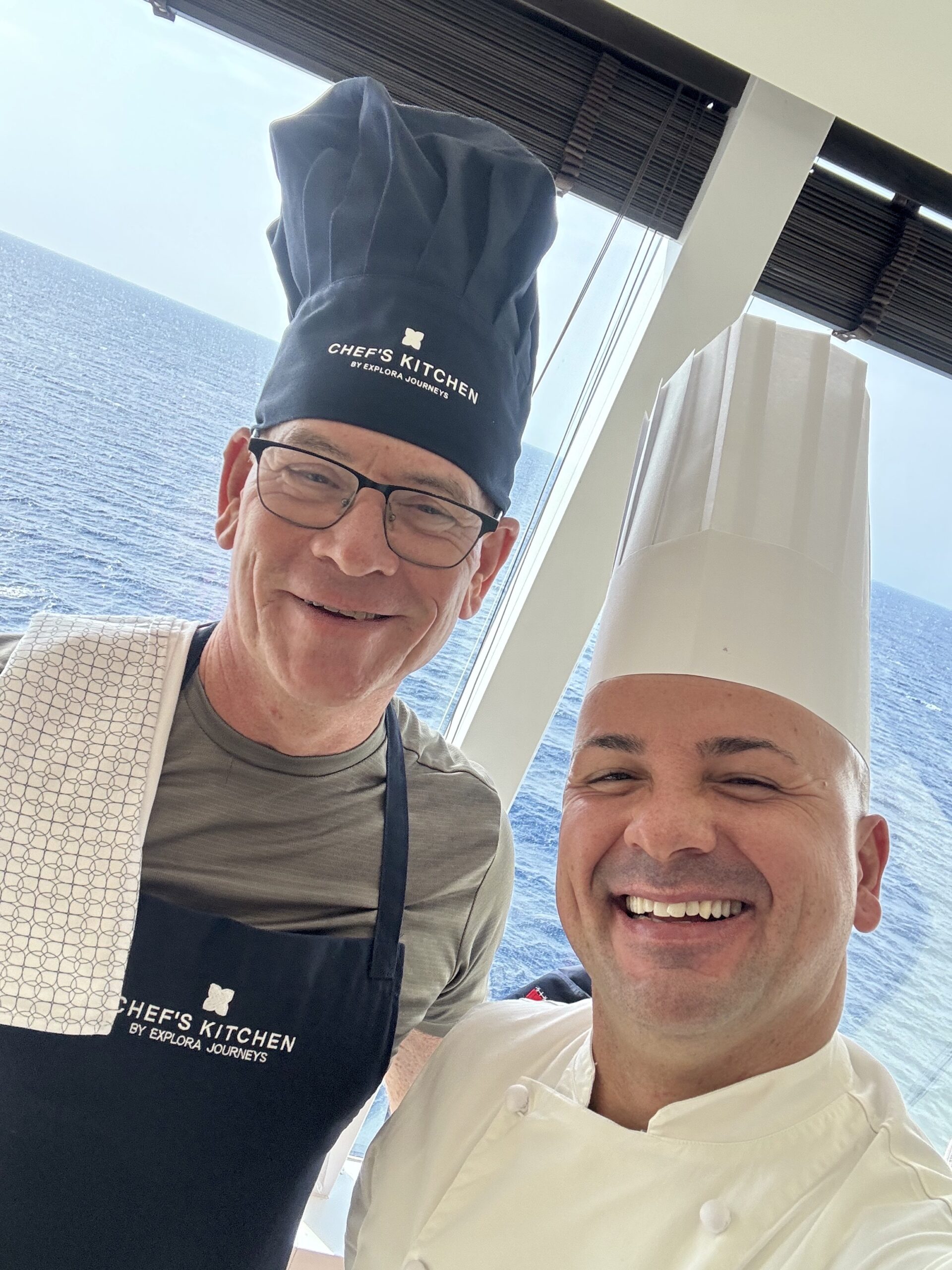 Two chefs posing for a selfie on a ship, one wearing a tall Chef's Kitchen hat and glasses with the sea behind them in the window.