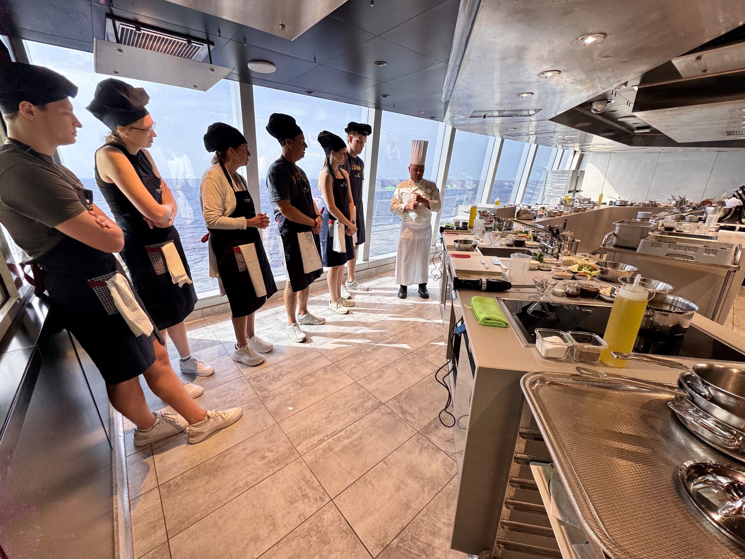 Line of aspiring chefs in black aprons and hats watch a chef in white uniform demonstration in a stainless steel galley with large windows over the water