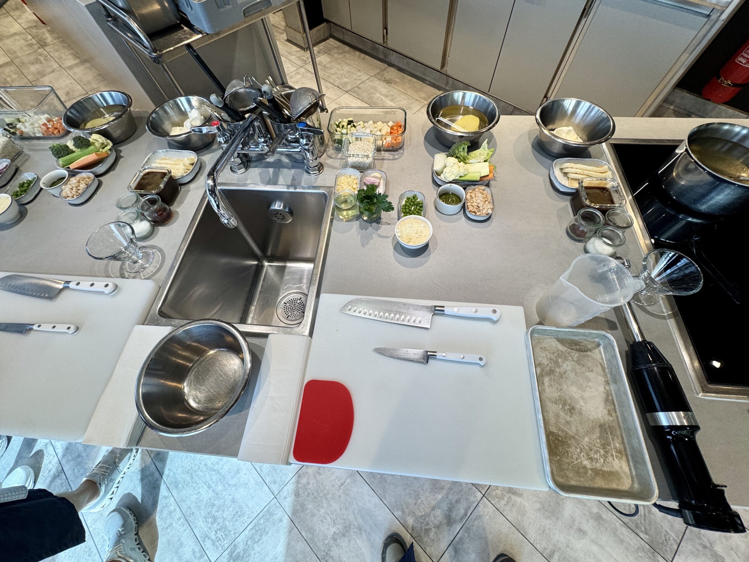 Top-down view of a kitchen prep counter with bowls of vegetables, sauces, herbs, and knives around a sink and cutting boards.