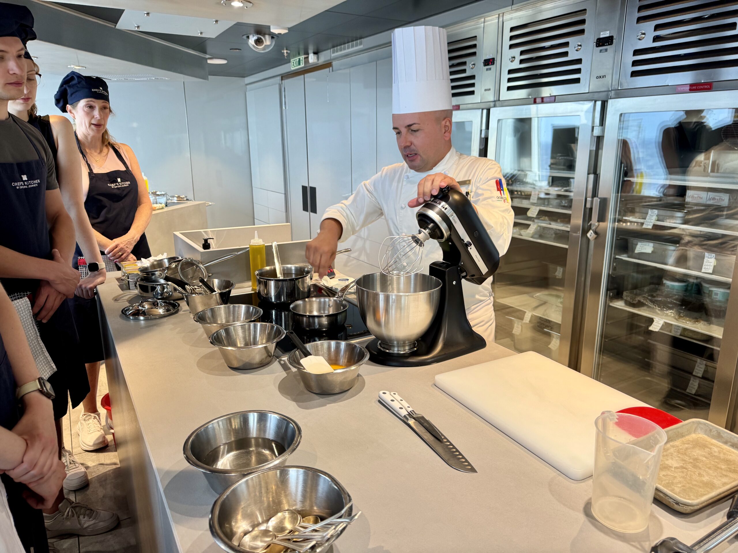 Chef in white uniform demonstrates with a stand mixer at a stainless steel prep counter while two assistants watch in a modern kitchen class.