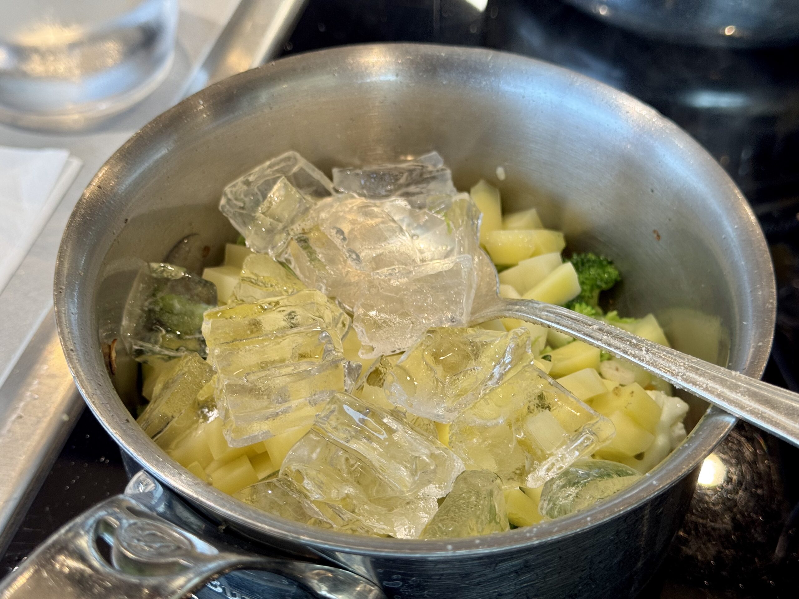 Stainless steel pot filled with ice cubes and diced potatoes, with broccoli visible beneath the ice.