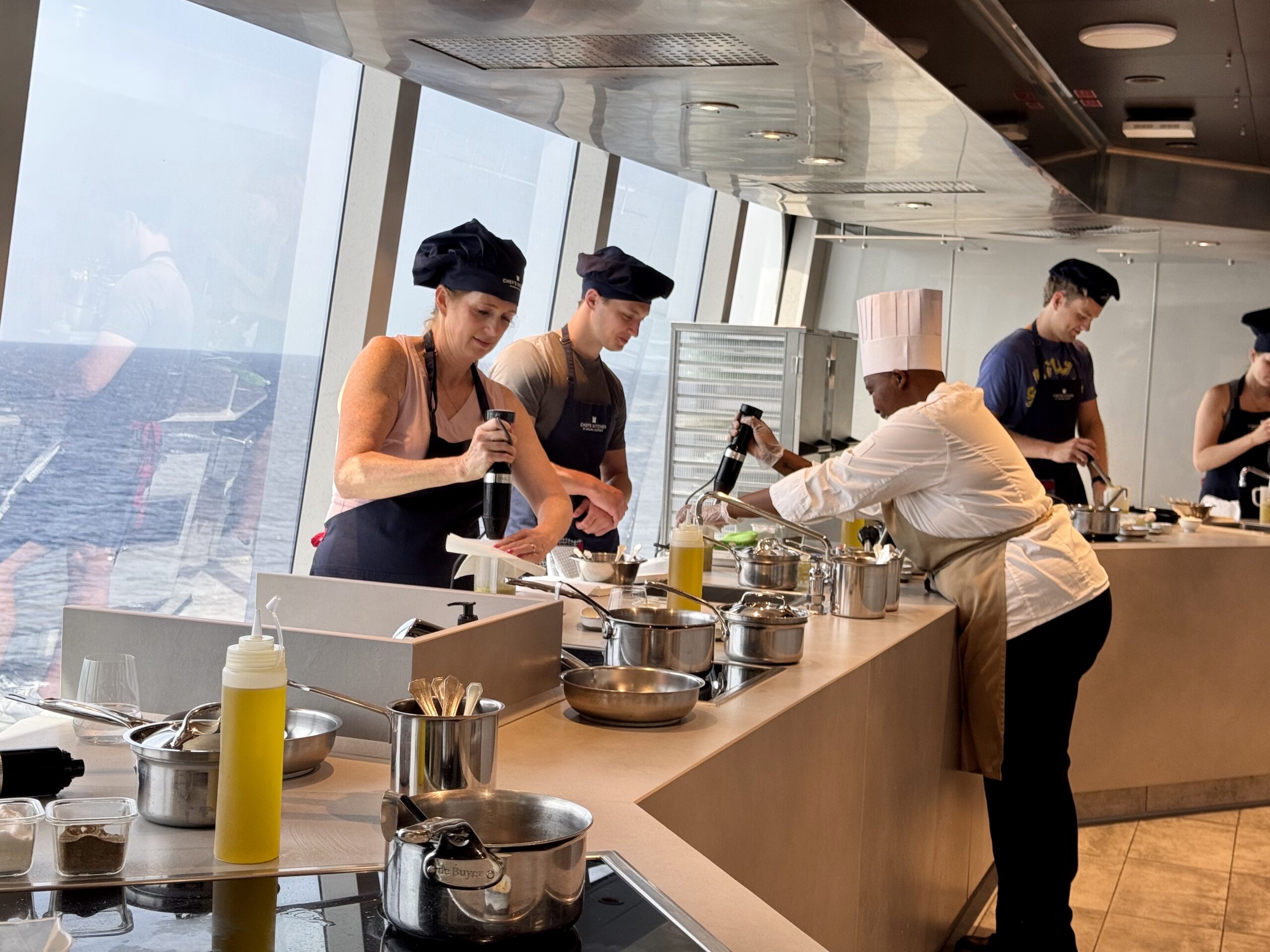 Group of chefs cooking at a ship kitchen station, preparing food with stainless pots and squeeze bottles, ocean visible through windows behind them.
