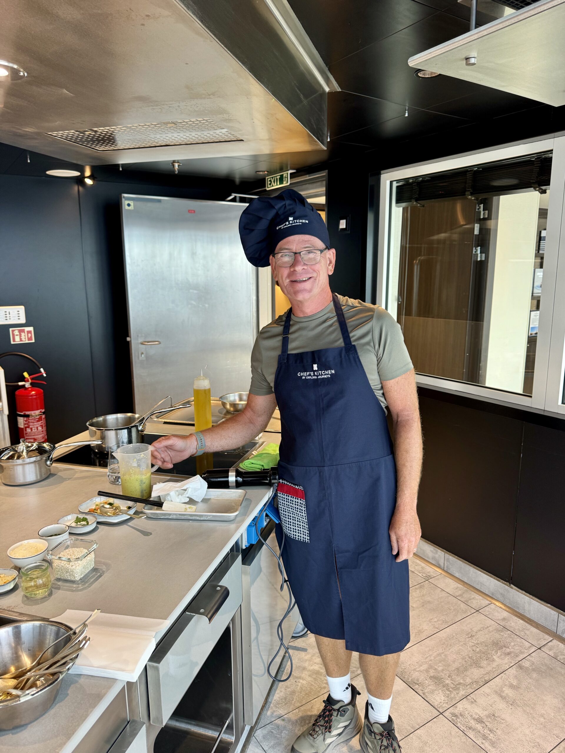 Male chef wearing a navy apron and hat stands at a stainless steel kitchen station with ingredients and utensils visible on the counter behind him.