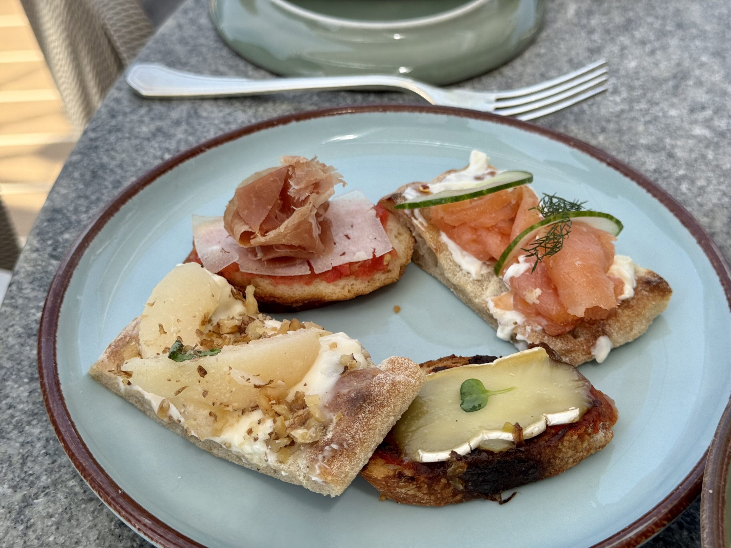 Assorted crostini on a blue-rimmed plate: salmon with cucumber and dill, prosciutto with cheese on crusty bread, and a lemon wedge nearby.