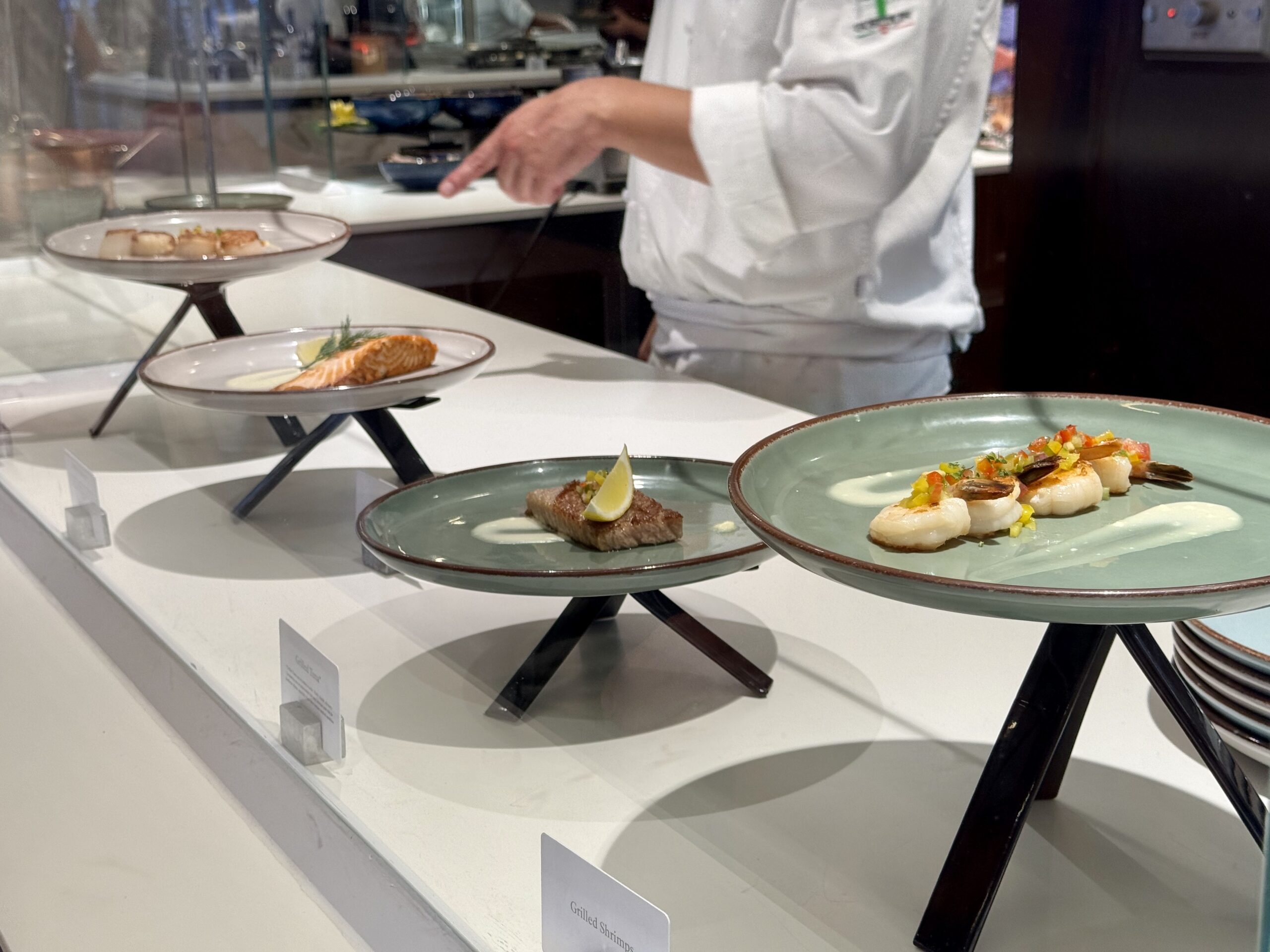 Chef in a white uniform arranging seafood dishes on a glass display counter with tiered plates in the foreground.