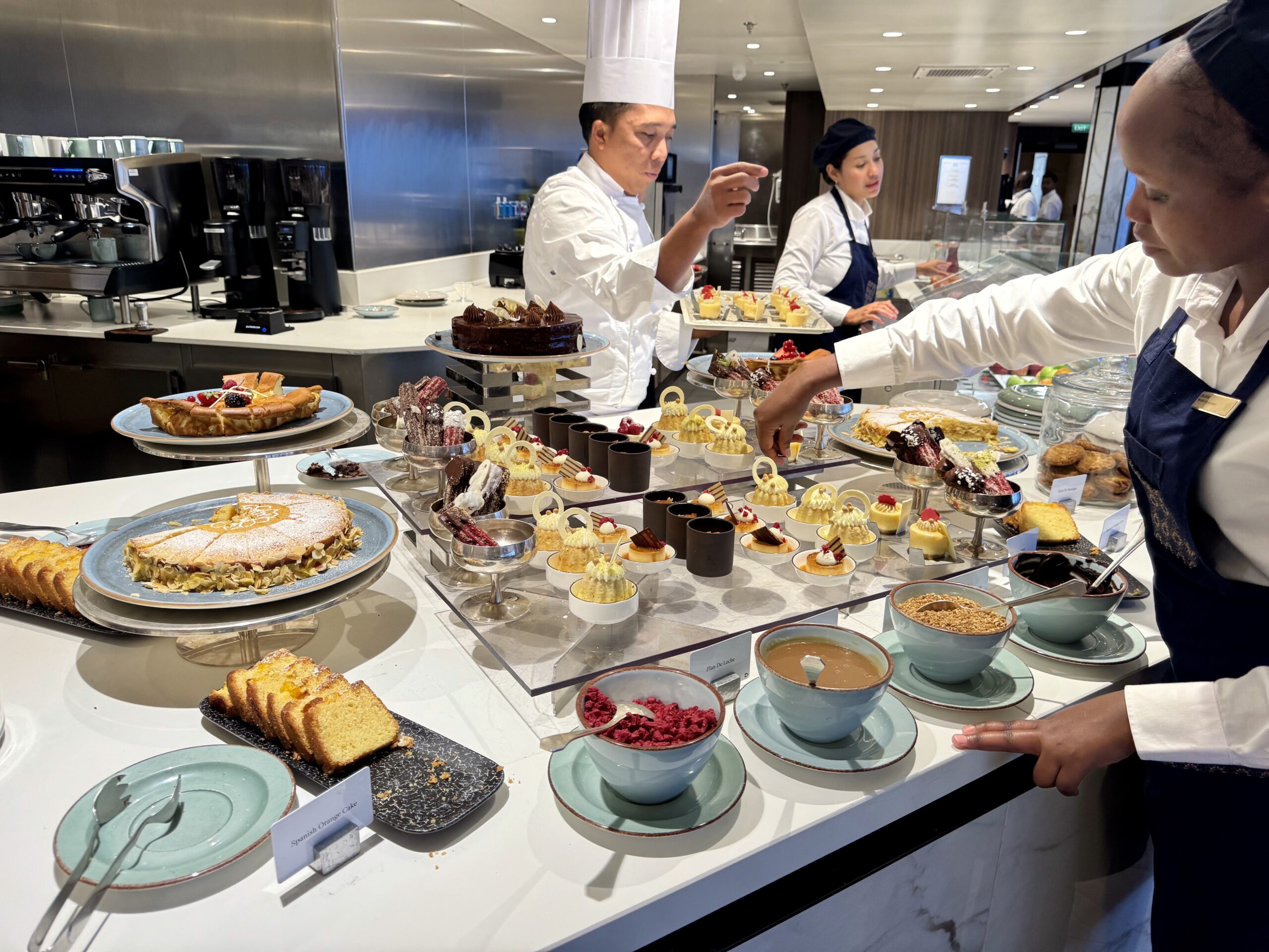 Pastry display in a cafe kitchen with assorted cakes, tarts, and sweets on glass stands in front of chefs at work.