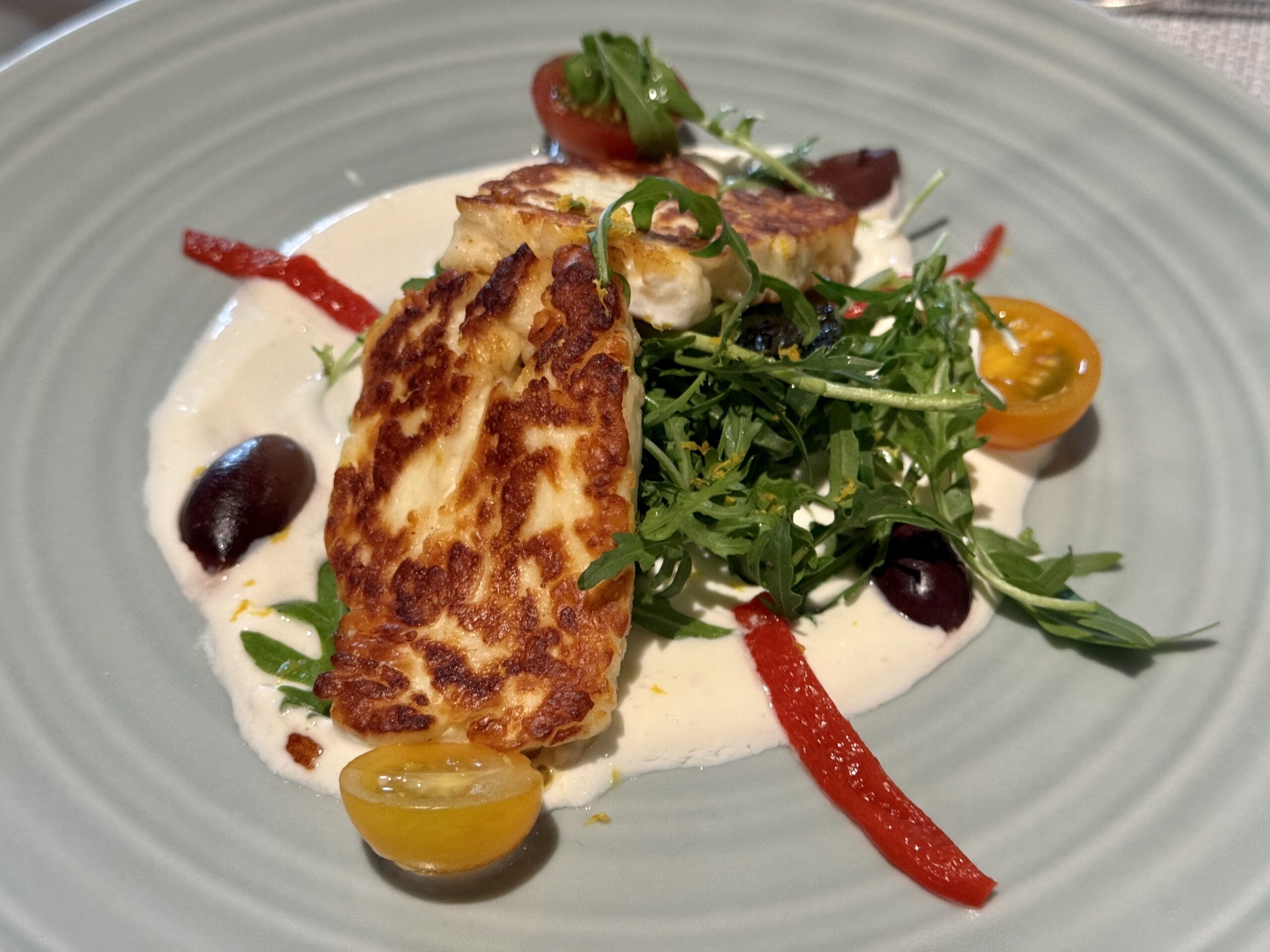 Seared fish fillet on a bed of greens with cream sauce, cherry tomatoes, olives, and a red sauce drizzle Replating details may vary for accessibility purposes.