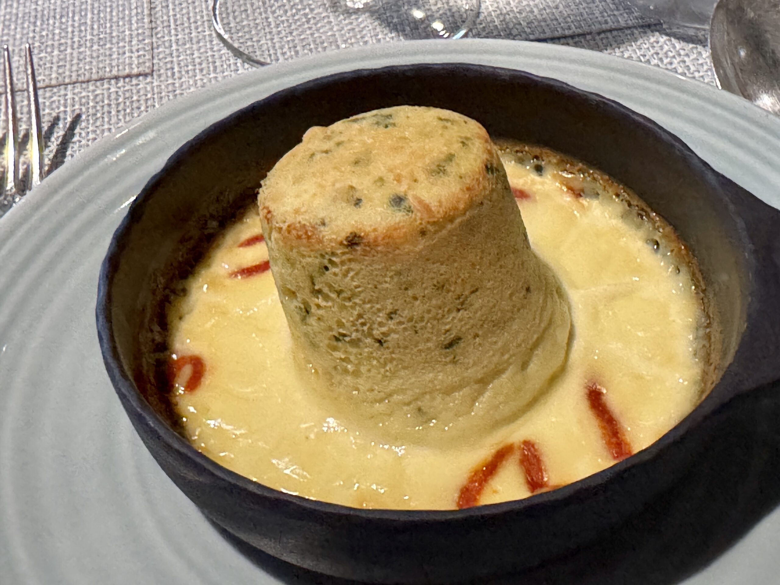 Creamy yellow soup in a small black skillet, topped with a round bread roll and red sauce streaks visible around the edge.