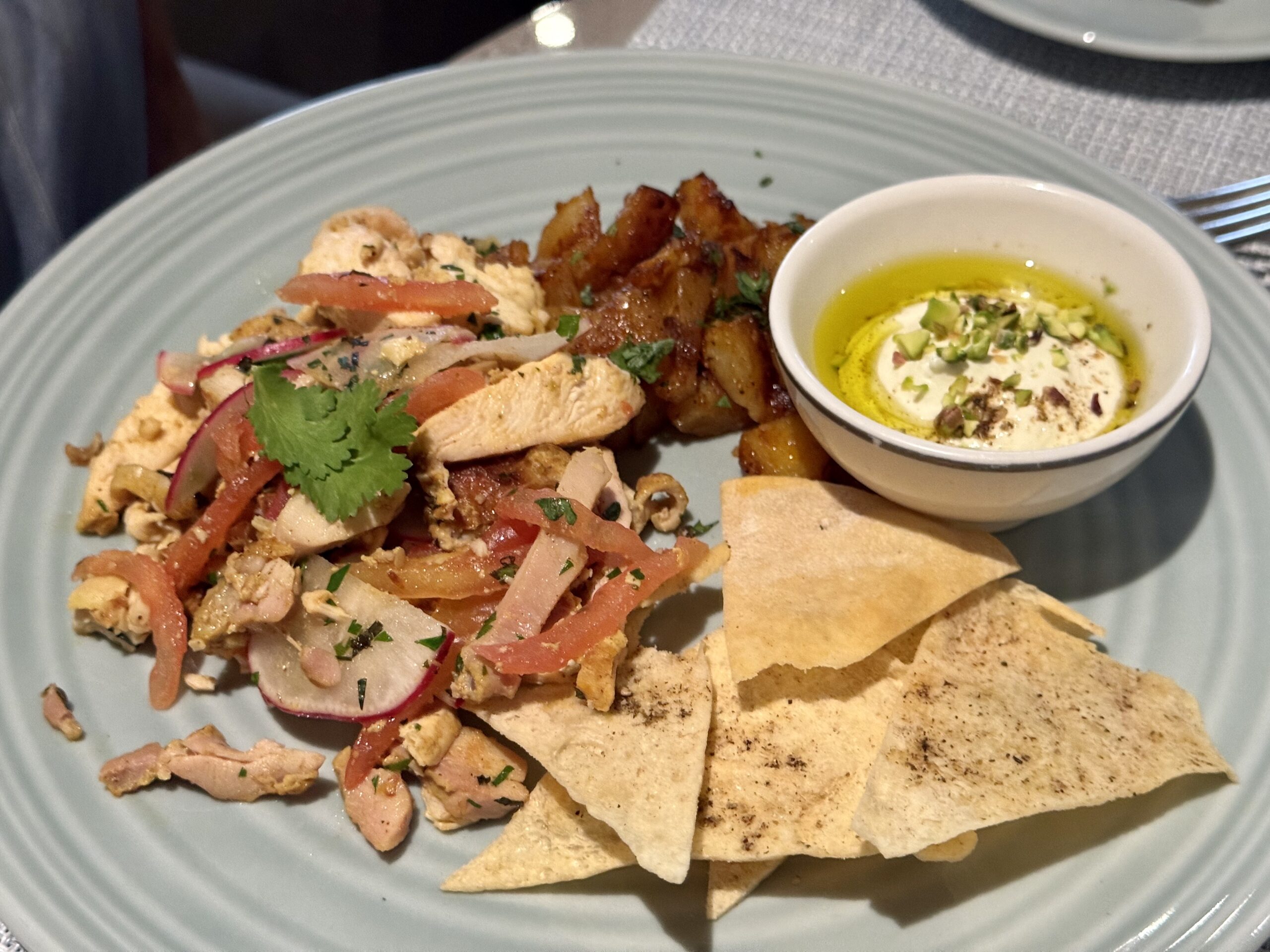 Sliced chicken with tomato and cilantro on a plate, served with tortilla chips and a small bowl of olive oil topped with yogurt and pistachios.