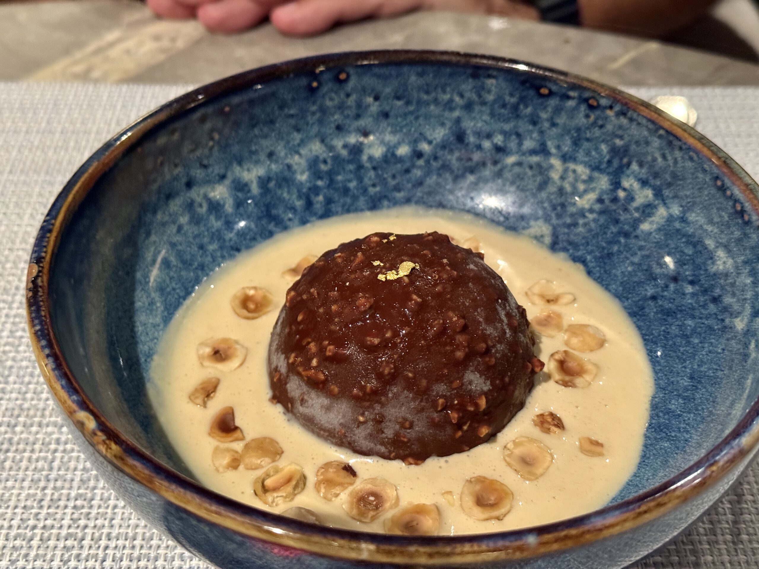 Blue ceramic bowl with a light brown creamy sauce, a dark chocolate dessert ball on top, sprinkled with edible gold leaf and nuts around the rim.