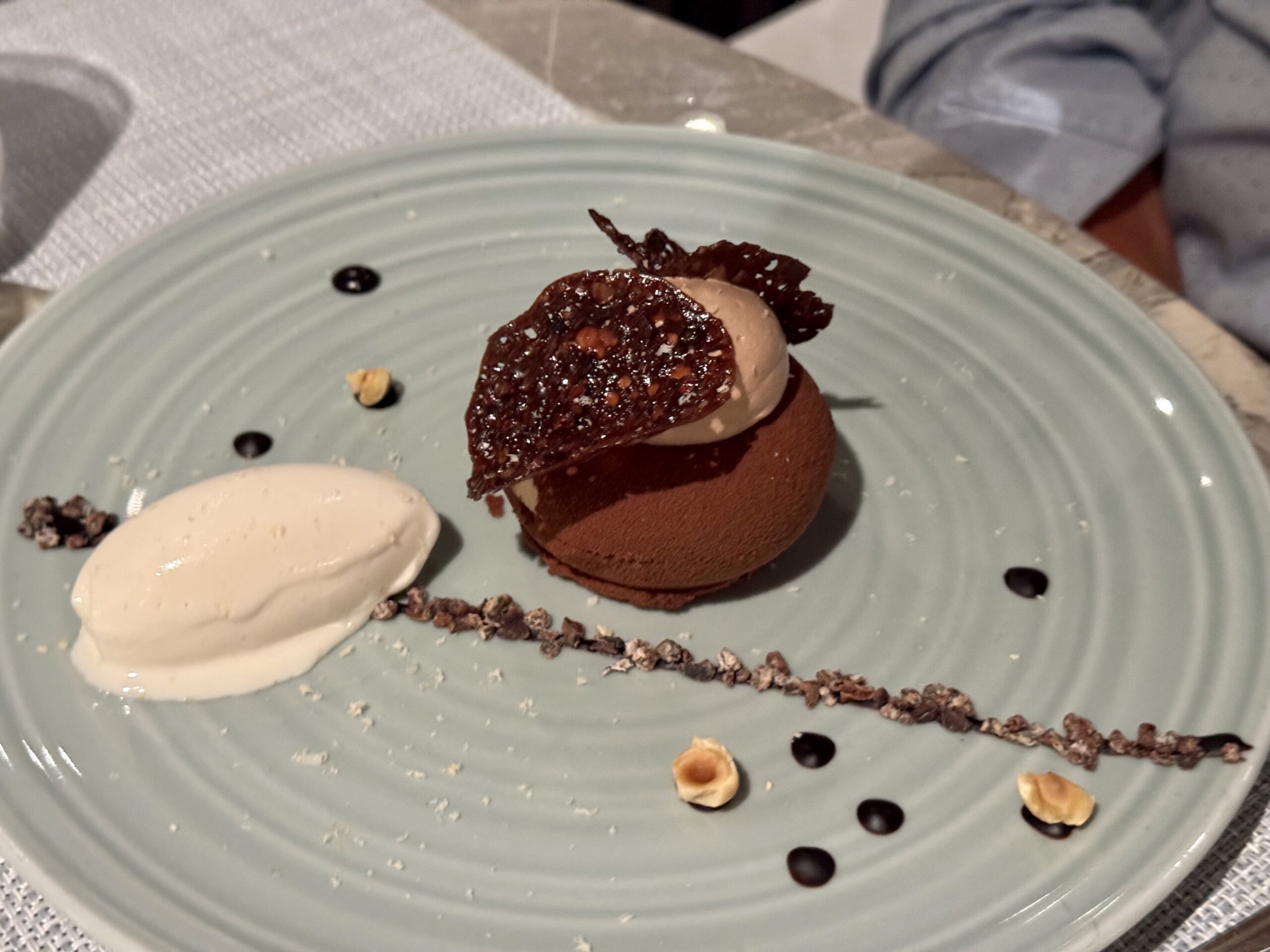 Chocolate cake bite with a crisp chocolate tuile, a scoop of vanilla ice cream, nuts, and chocolate drizzle on a pale blue plate.