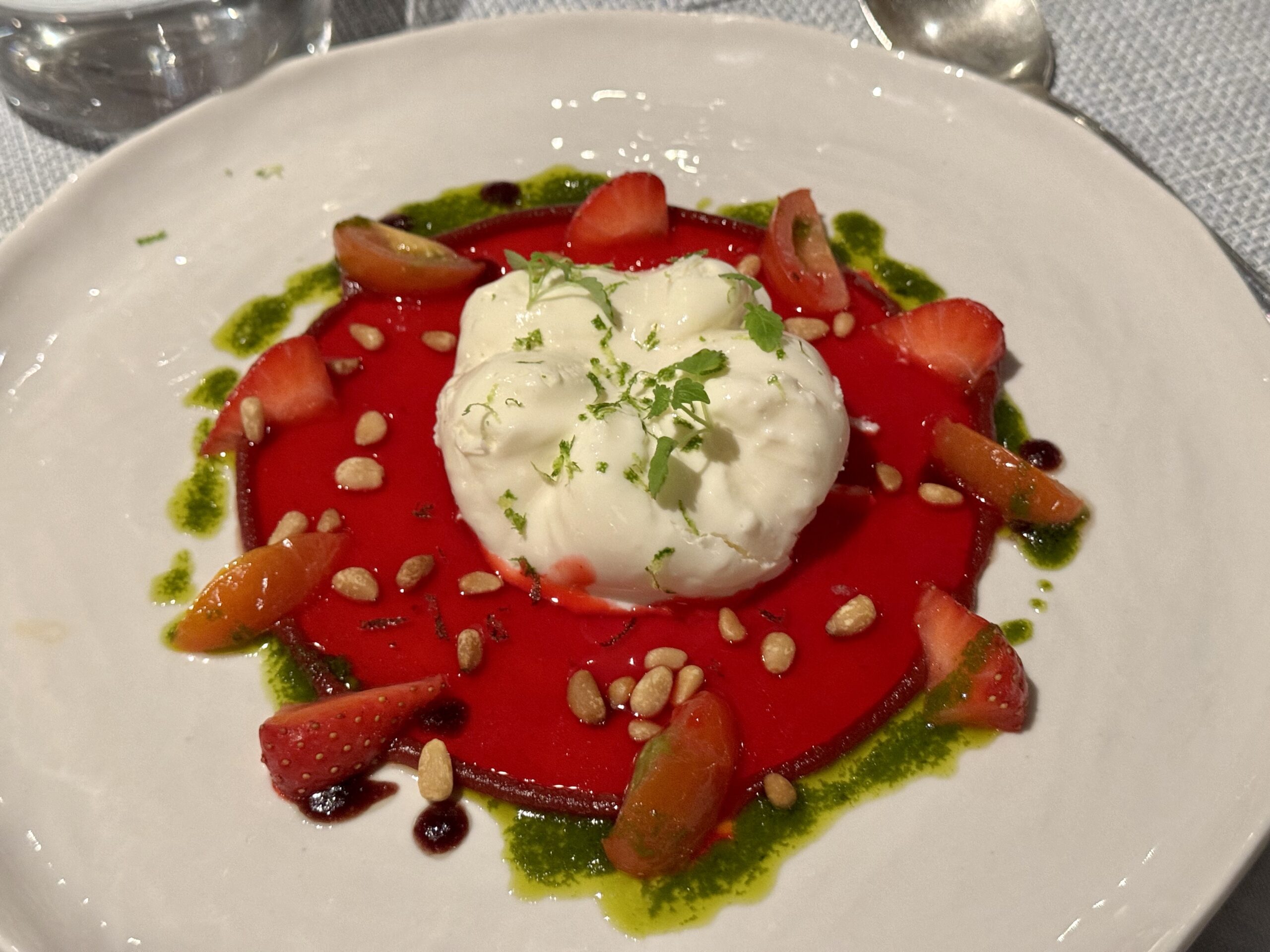 Burrata atop a red sauce with strawberry slices, green herb oil, pine nuts, and a mint garnish on a white plate.