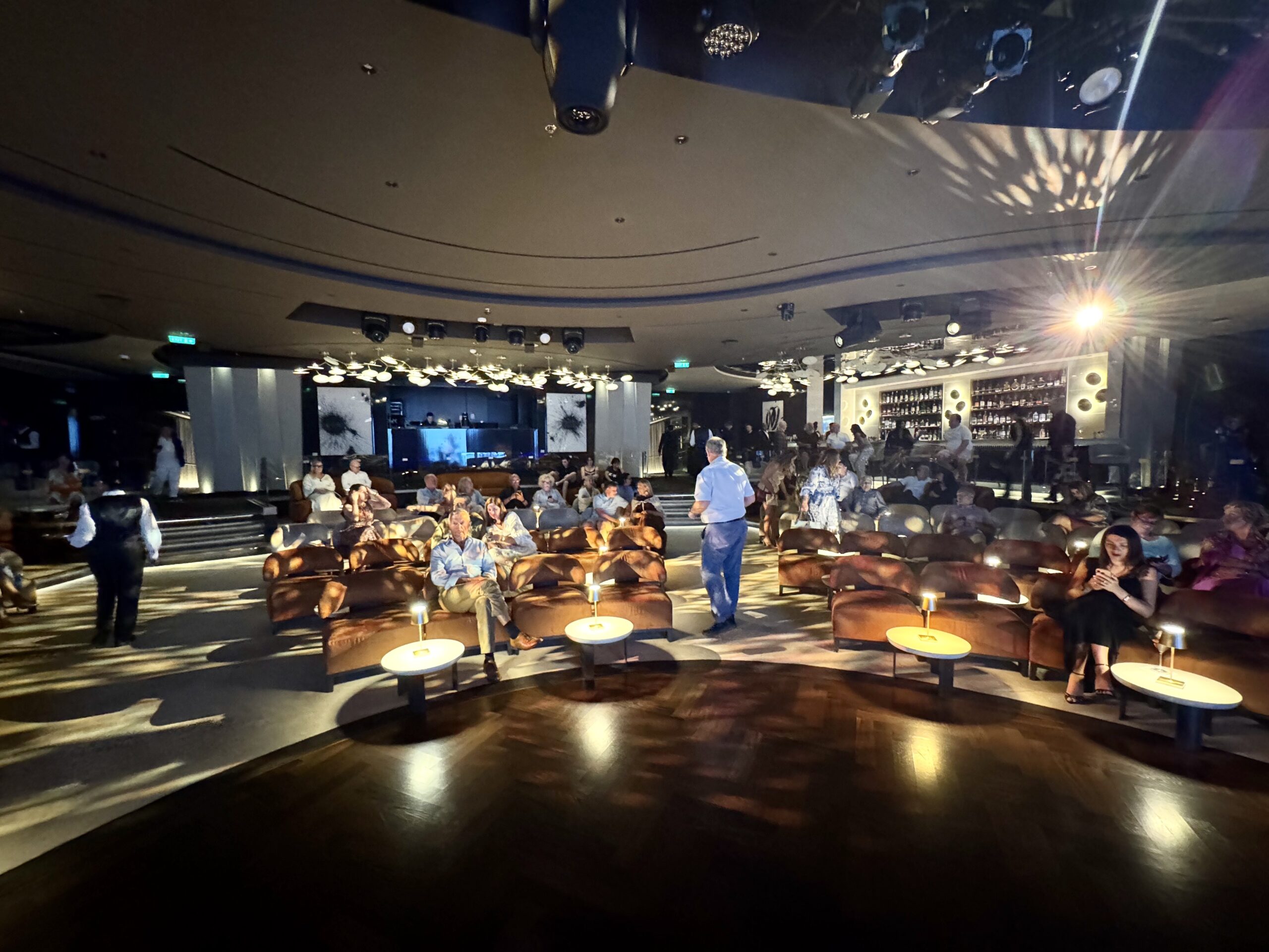 Dimly lit lounge with circular tables and brown leather chairs; people seated and chatting.