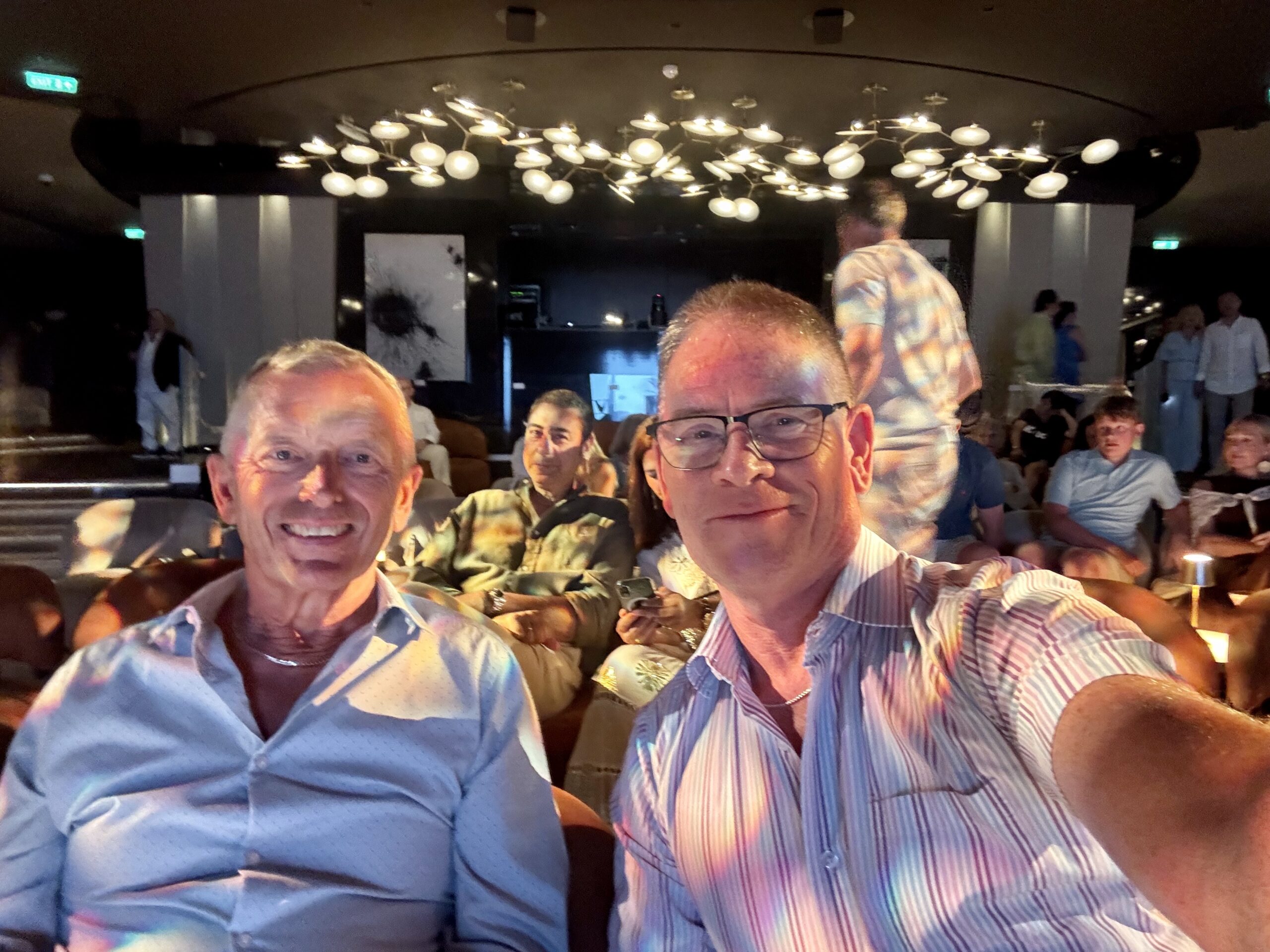 Two smiling men taking a selfie in a dimly lit lounge with a modern chandelier overhead and people seated in the background.