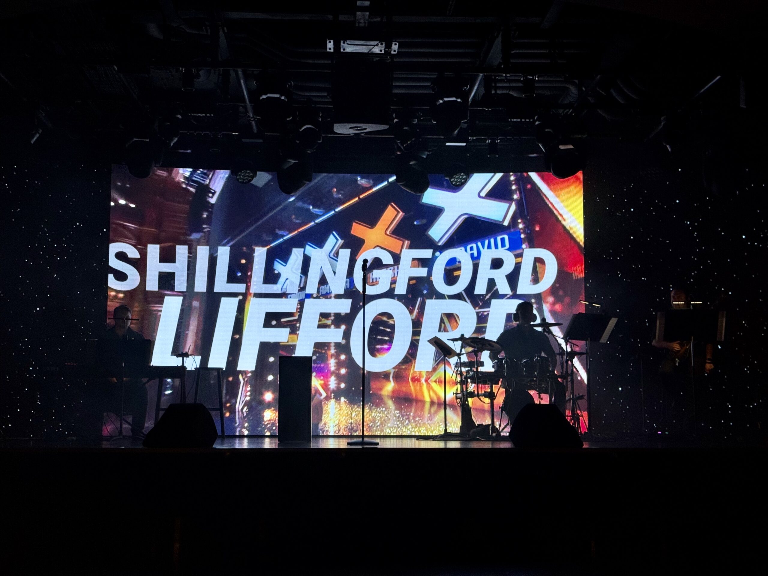Stage performance with a bright, multicolored screen showing bold white text (likely band name); silhouettes of musicians and instruments on a dark stage with a starry backdrop behind them.