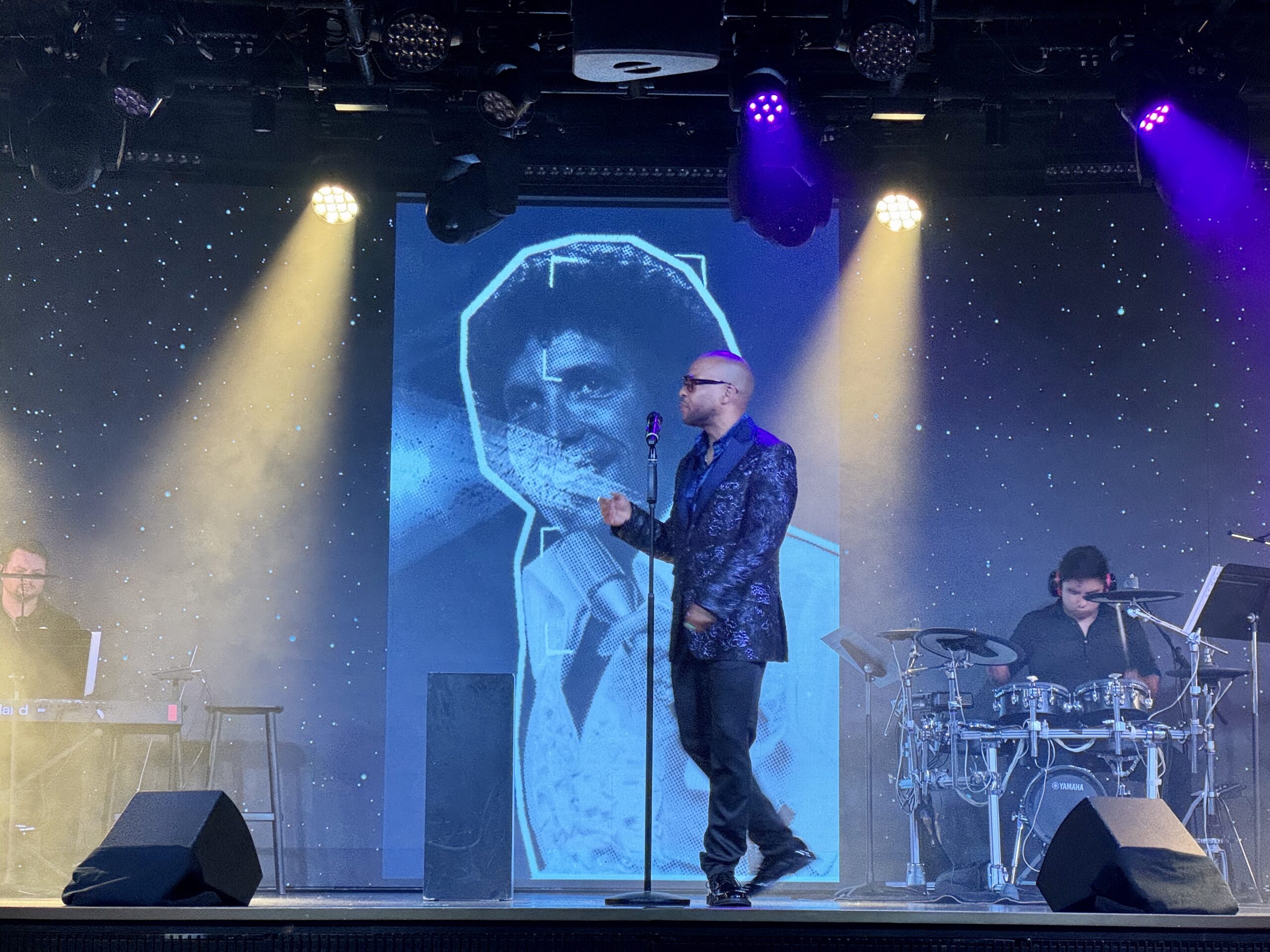 Male singer in dark suit performing on stage with microphones, blue neon backdrop, and a drummer to the right.