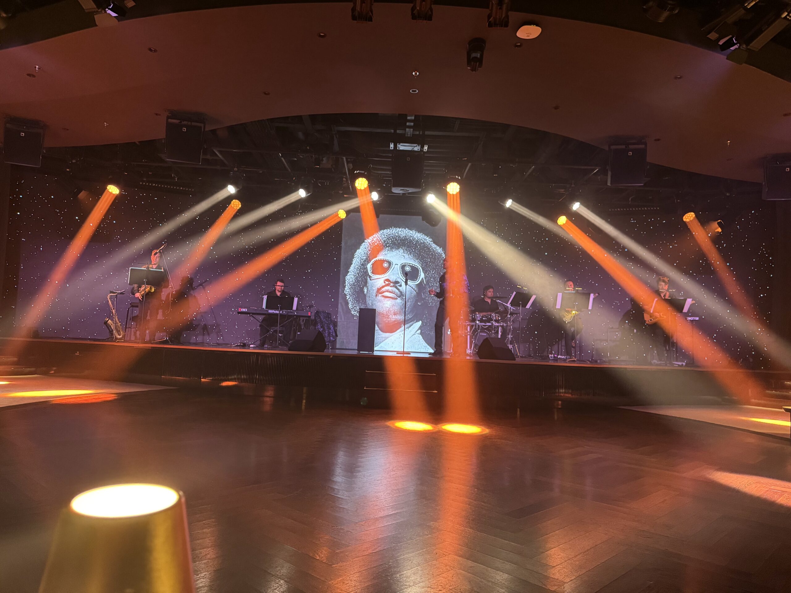 Concert stage with musicians playing keyboards, saxophone and guitar under orange and white spotlights against a starry backdrop; a large portrait image is projected on the screen behind them.