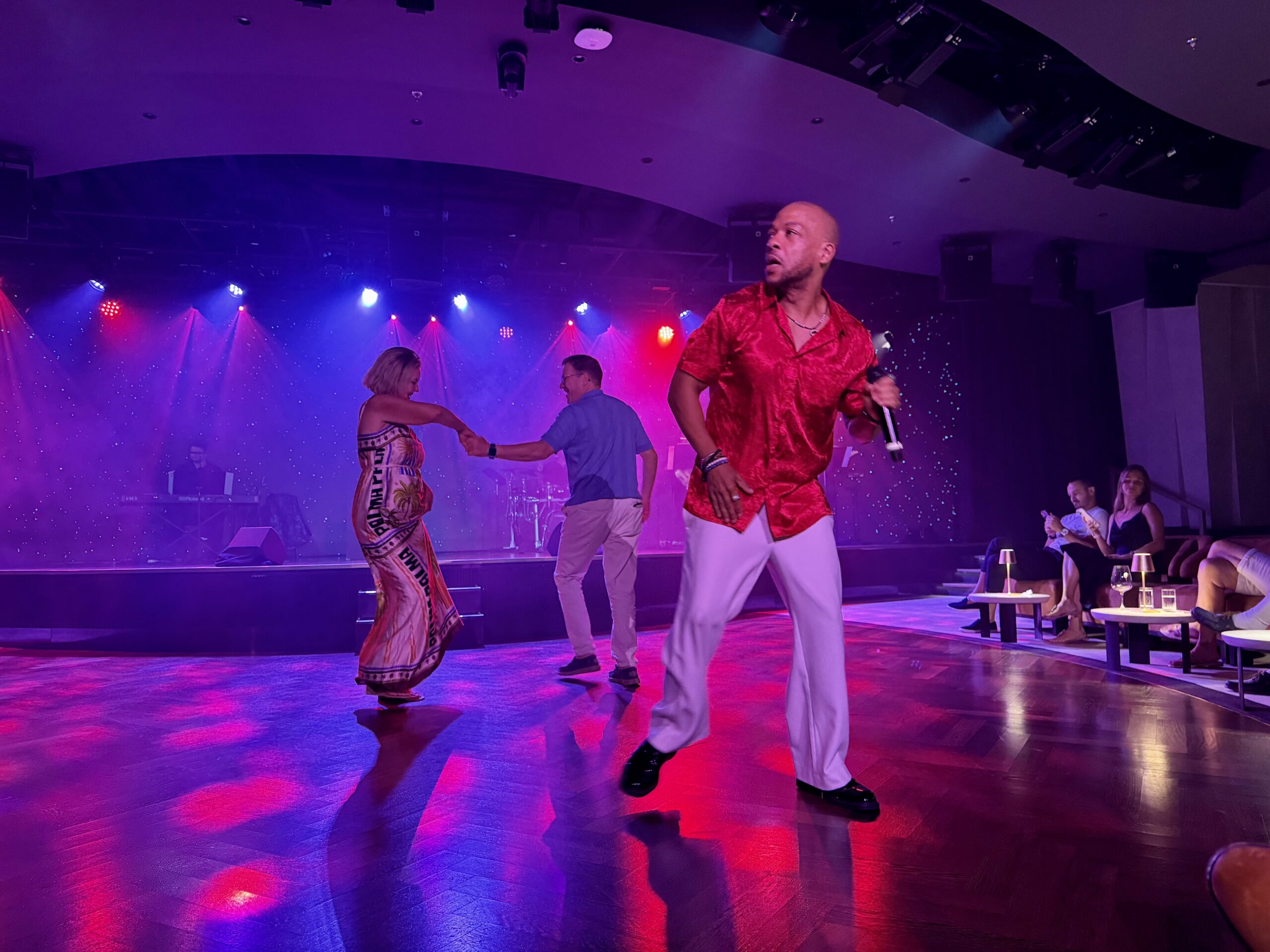 Stage performance with dancers under purple and red lights, a man in a red shirt front and center holding a microphone while others dance.