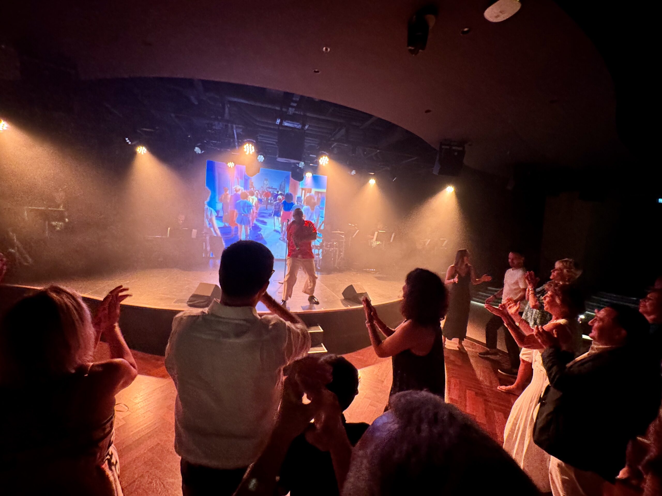 Audience in a circular formation around a lit stage with dancers and a large screen backdrop