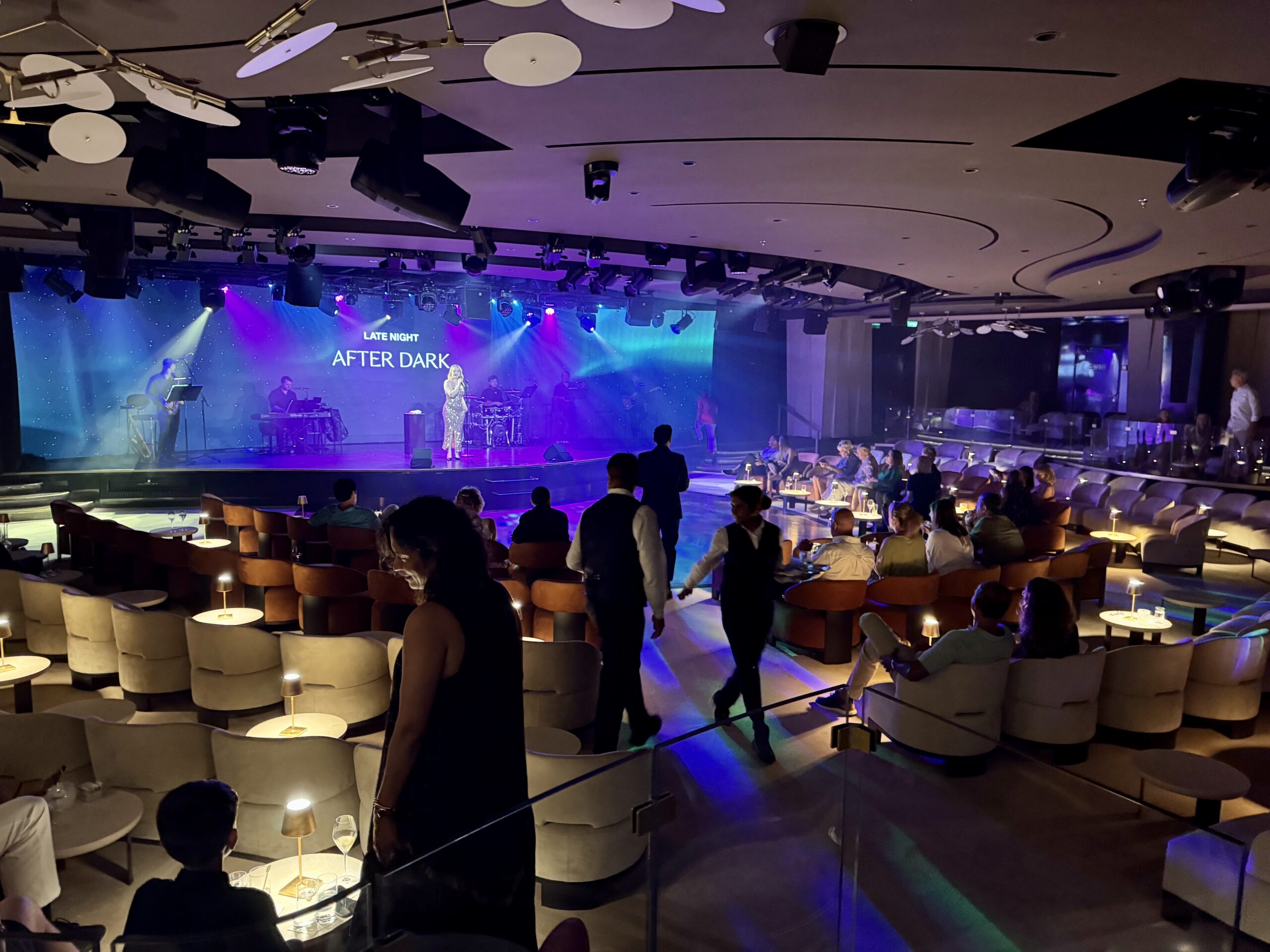 Stage performance with a singer under blue/purple lights; a band is playing on a lit backdrop that reads 'Late Night After Dark' while audience sits at tables in a lounge area.