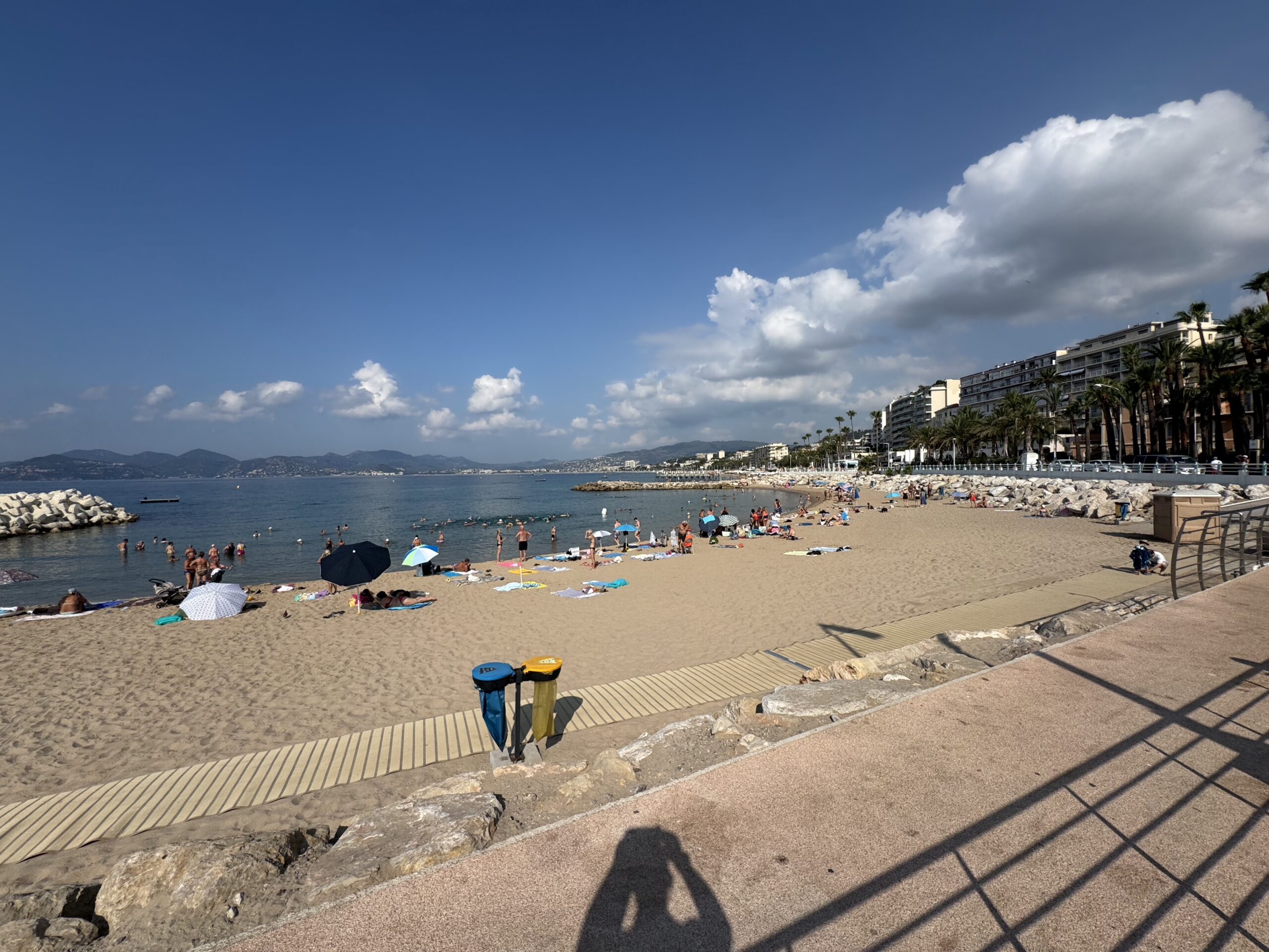 Busy beach scene with people sunbathing under umbrellas along a sandy shore, calm blue sea to the left and a row of palm-lined buildings on the right under a clear sky.