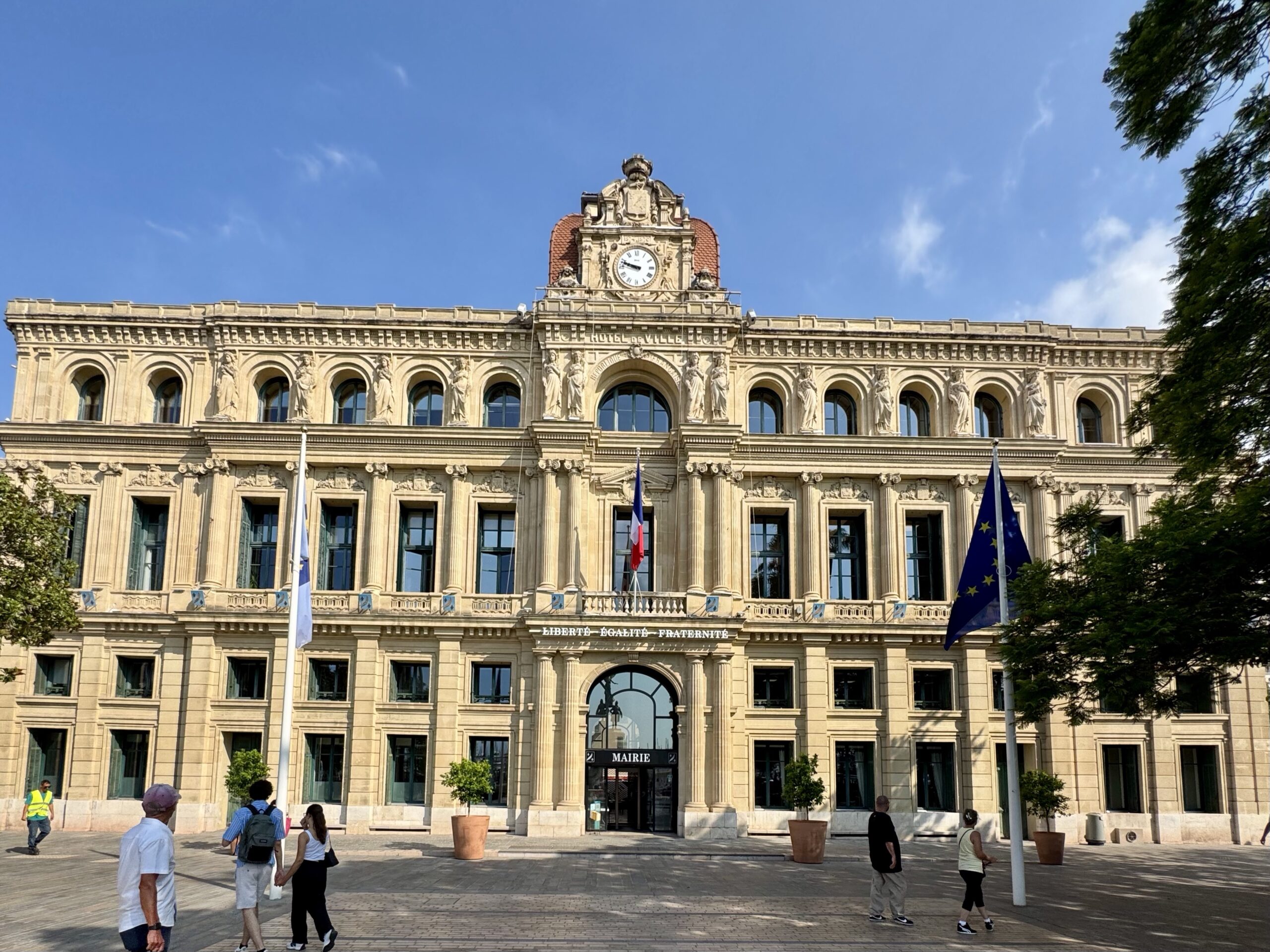 Beige historic town hall with a central clock tower, arched windows, statues along the façade, and flagpoles in front.