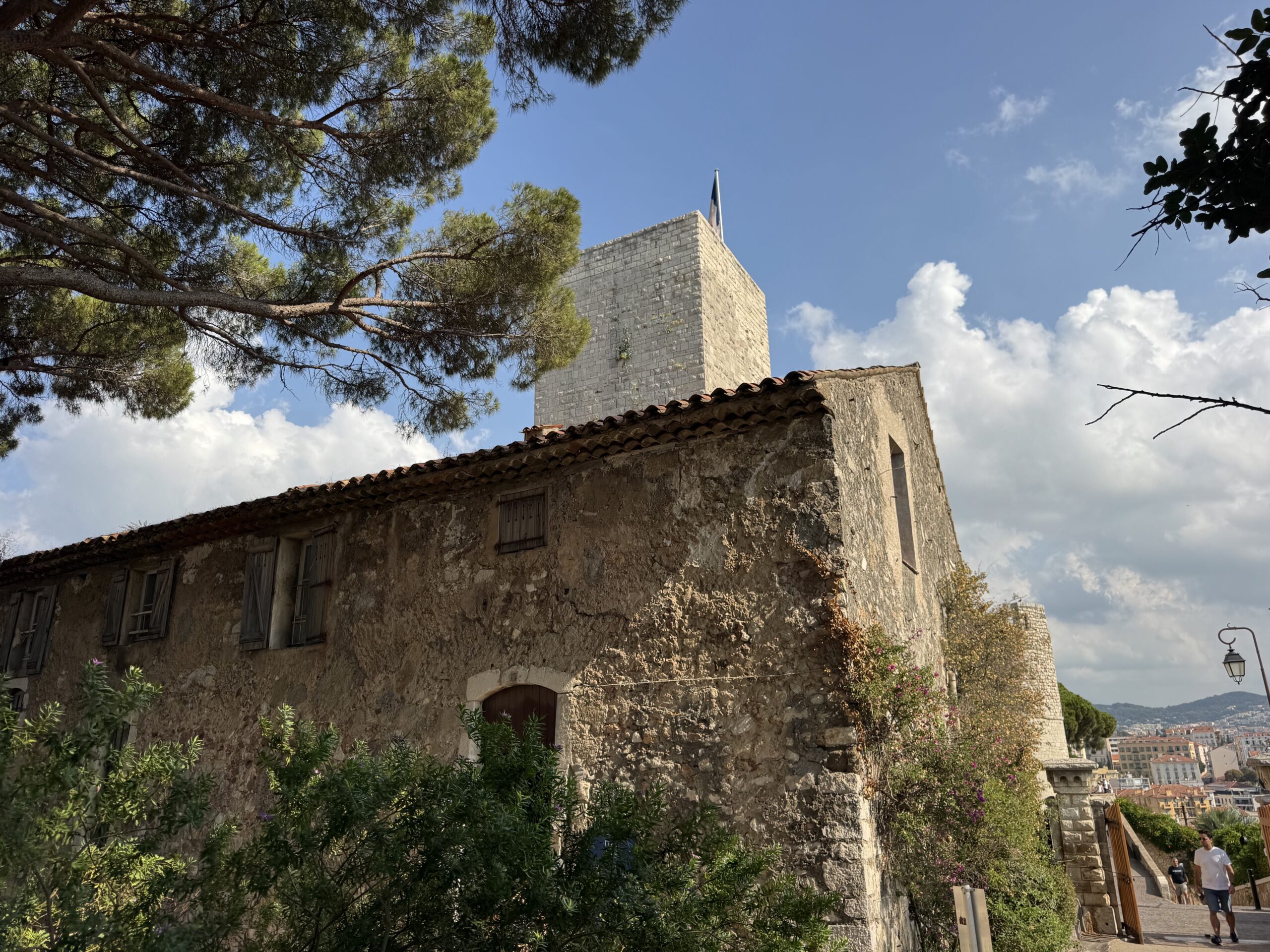 Stone building with a square tower and flag atop, framed by pine branches against a blue sky.
