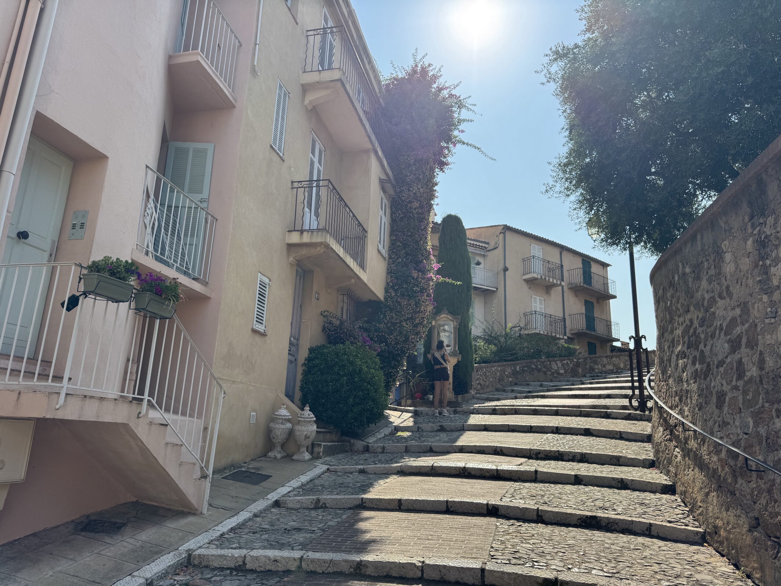 A sunlit narrow stairway between pastel buildings with balconies and a stone wall on the right, under a bright blue sky.