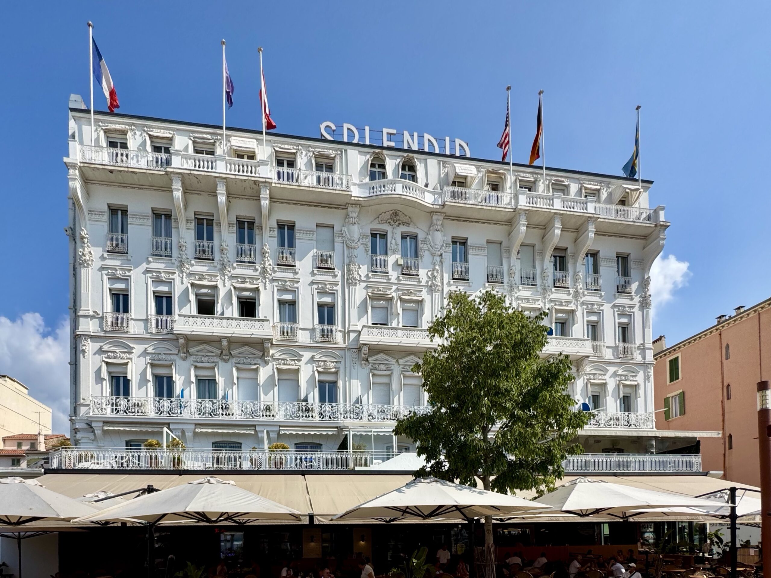 White ornate, multi-story building with decorative balconies and flagpoles atop, set against a clear blue sky, suggesting a grand hotel or historic edifice with flags flying.