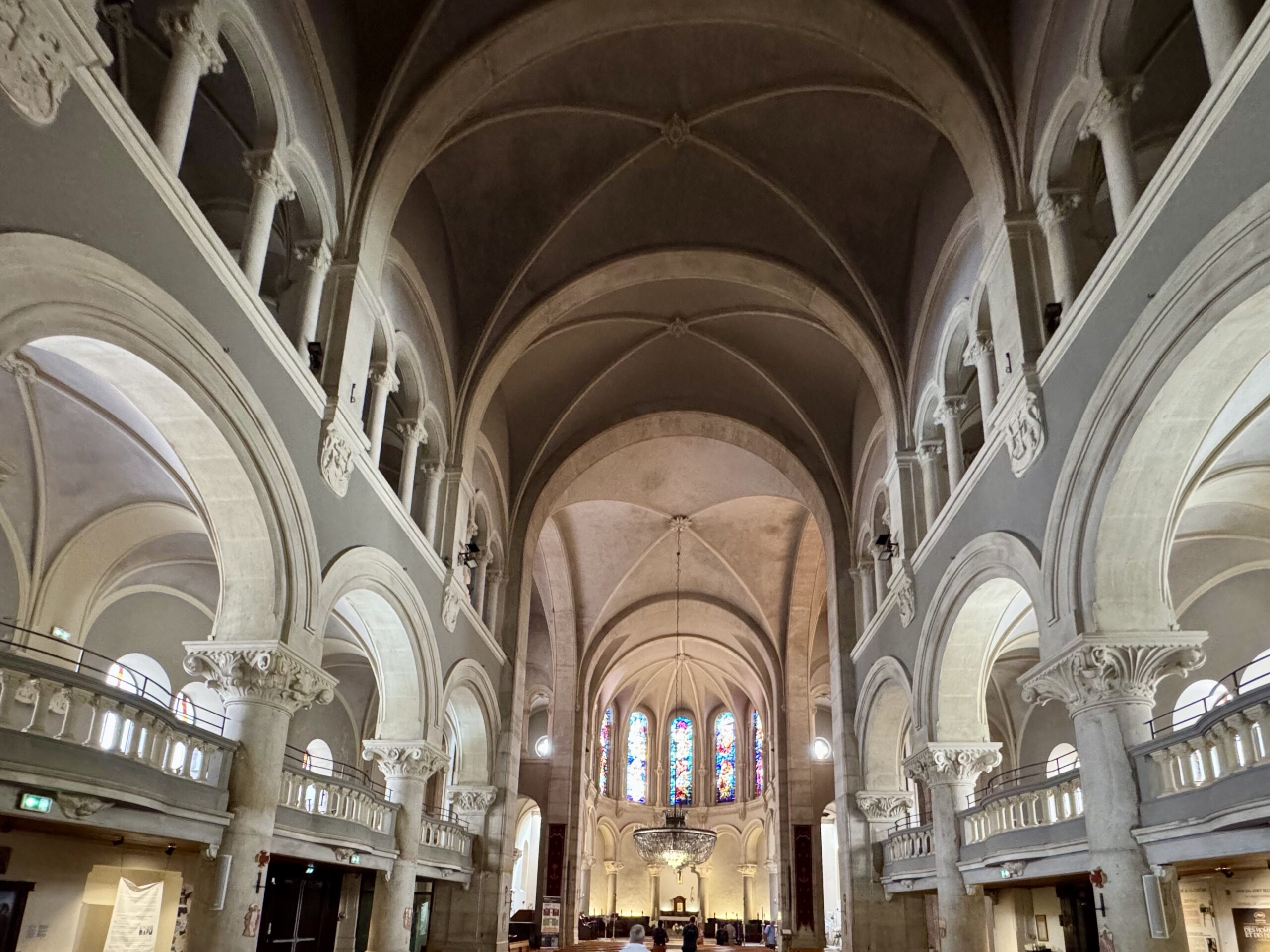 Interior of a grand cathedral with tall vaulted arches, ornate columns, and a stained-glass window at the far end.