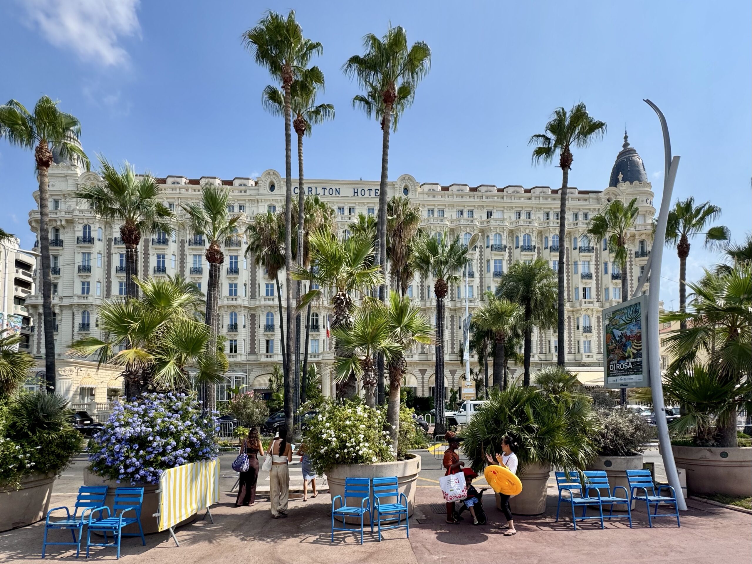 Elegant hotel facade with ornate windows and arches, framed by tall palm trees in a sunny plaza.